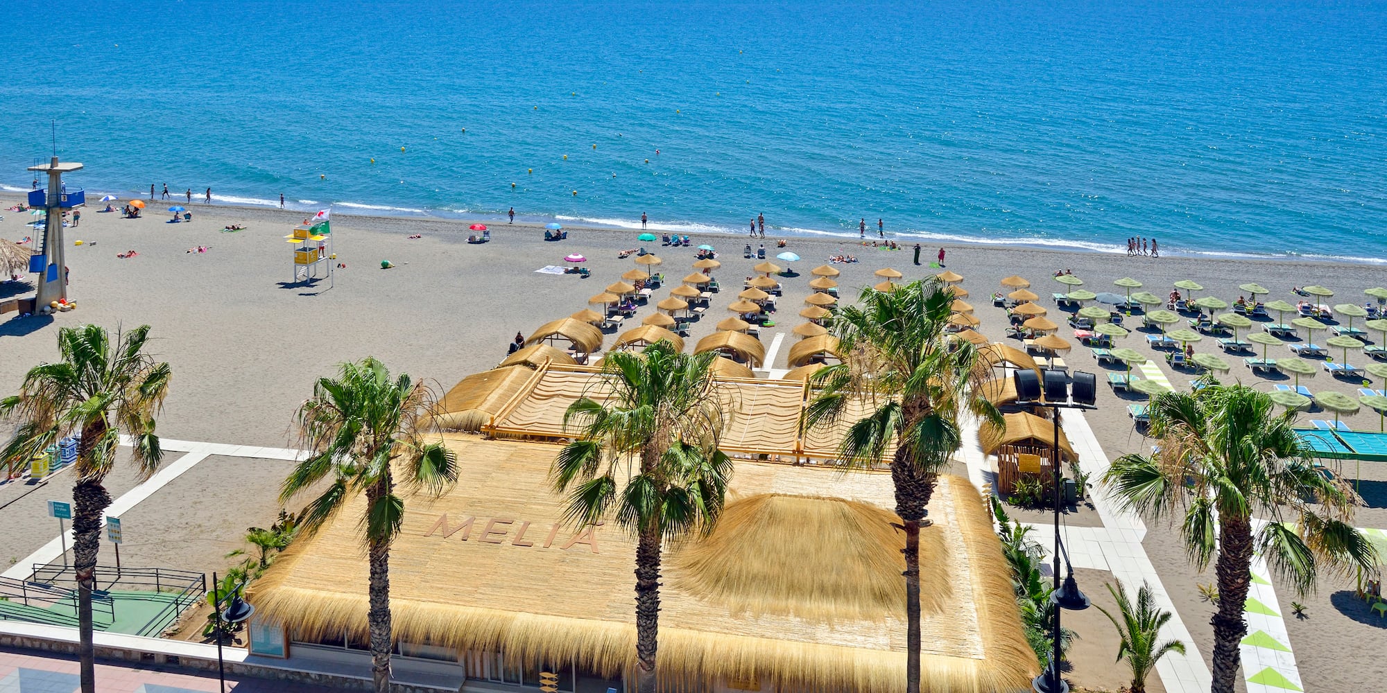 Sunny Melia beach club with palm trees, blue sea, and numerous beach umbrellas.