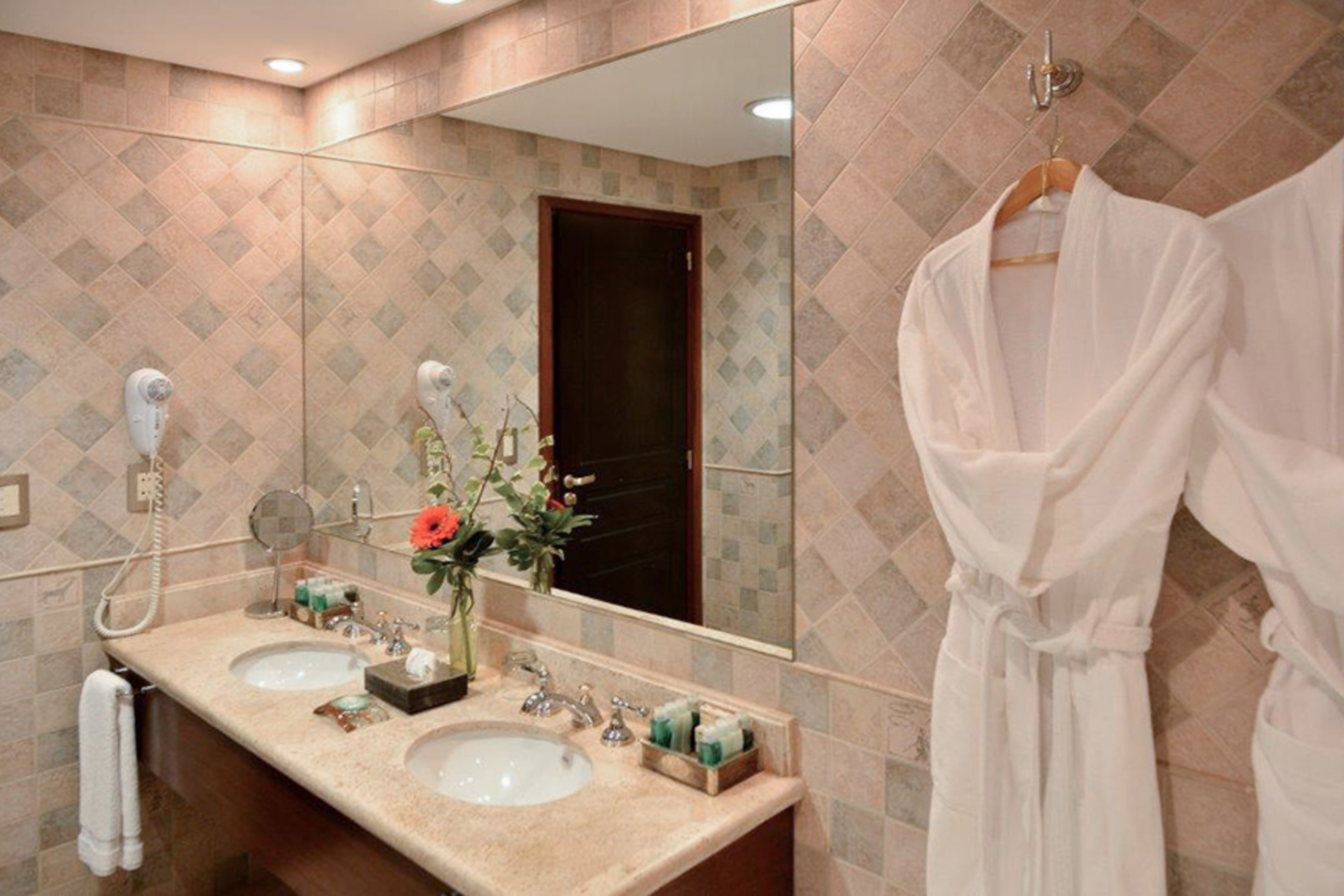 Hotel bathroom with double sinks, mirror, tiled walls, and white bathrobes.