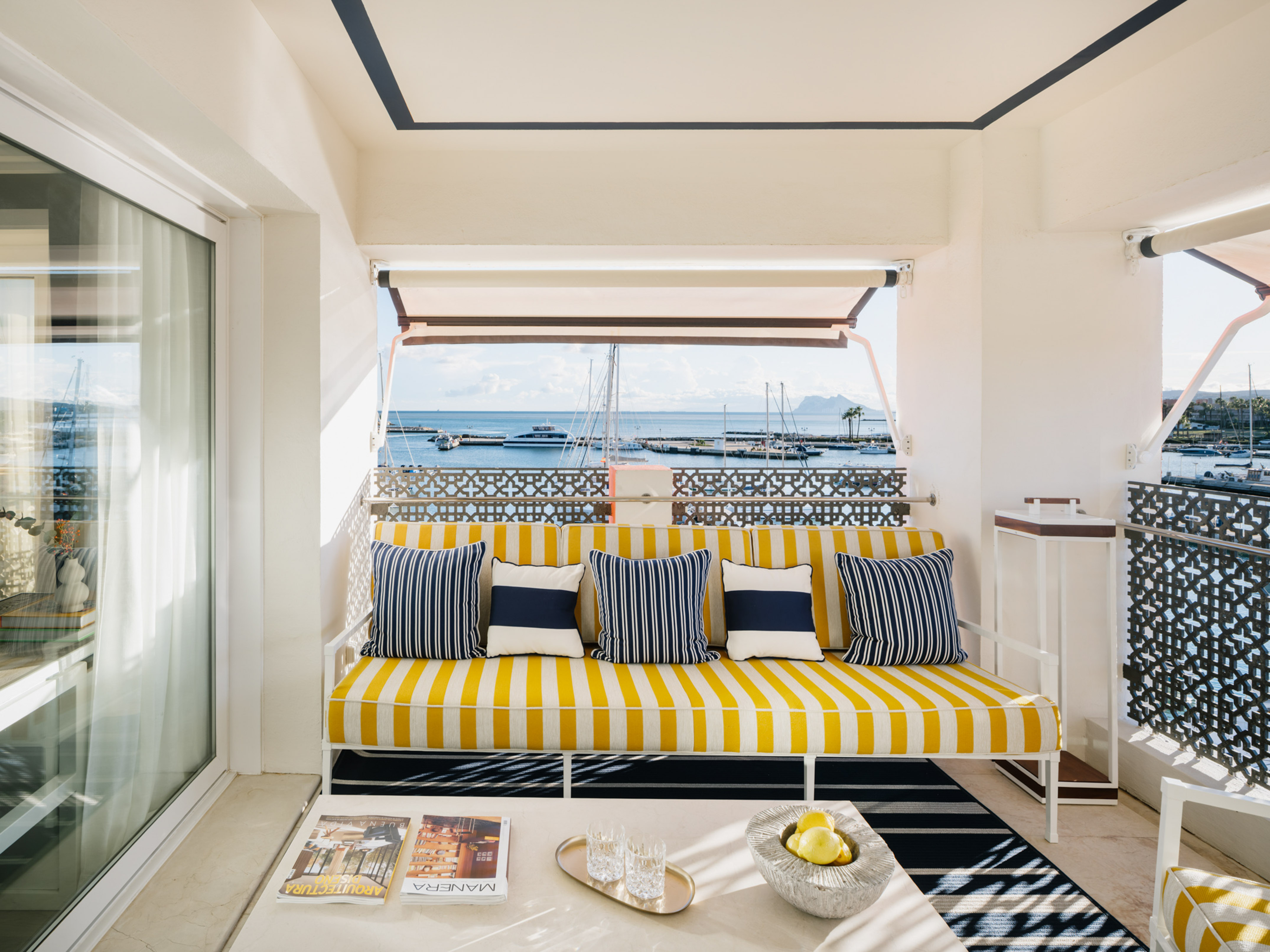Sunny balcony with yellow striped sofa, nautical cushions, and marina view.