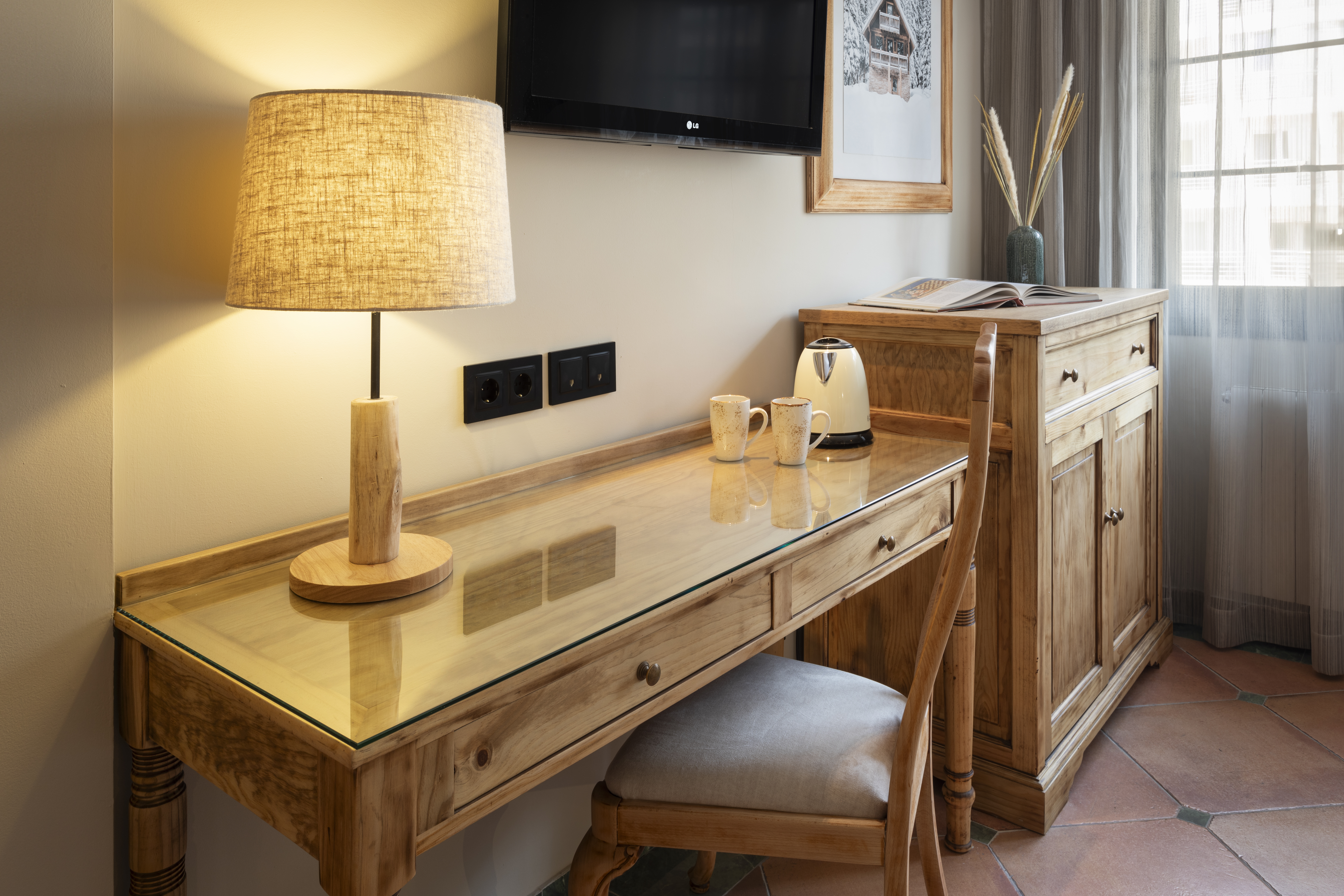 Hotel room workspace with natural wood desk, lamp, kettle, mugs, and TV.