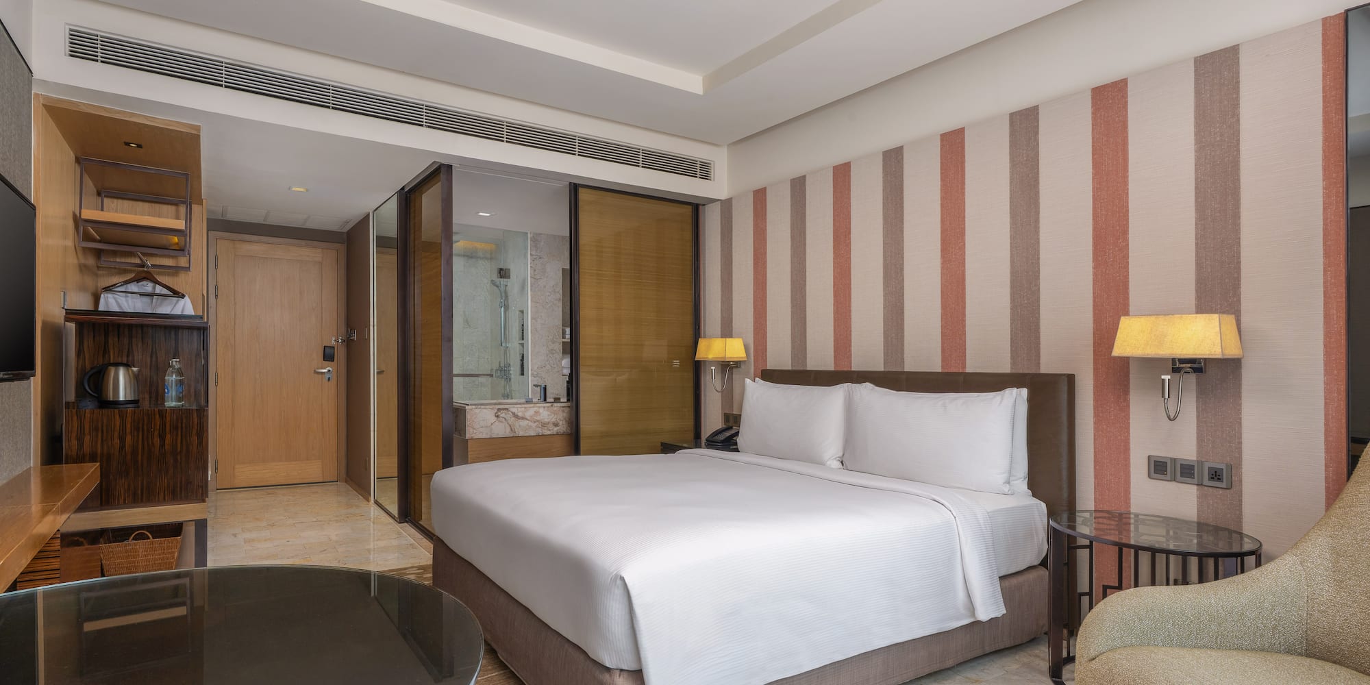 Modern hotel room with a king bed, striped accent wall, and glass-partitioned bathroom. - Guest Room - The Hotel Bangkok Sukhumvit 26 Affiliated by Meliá, Bangkok
