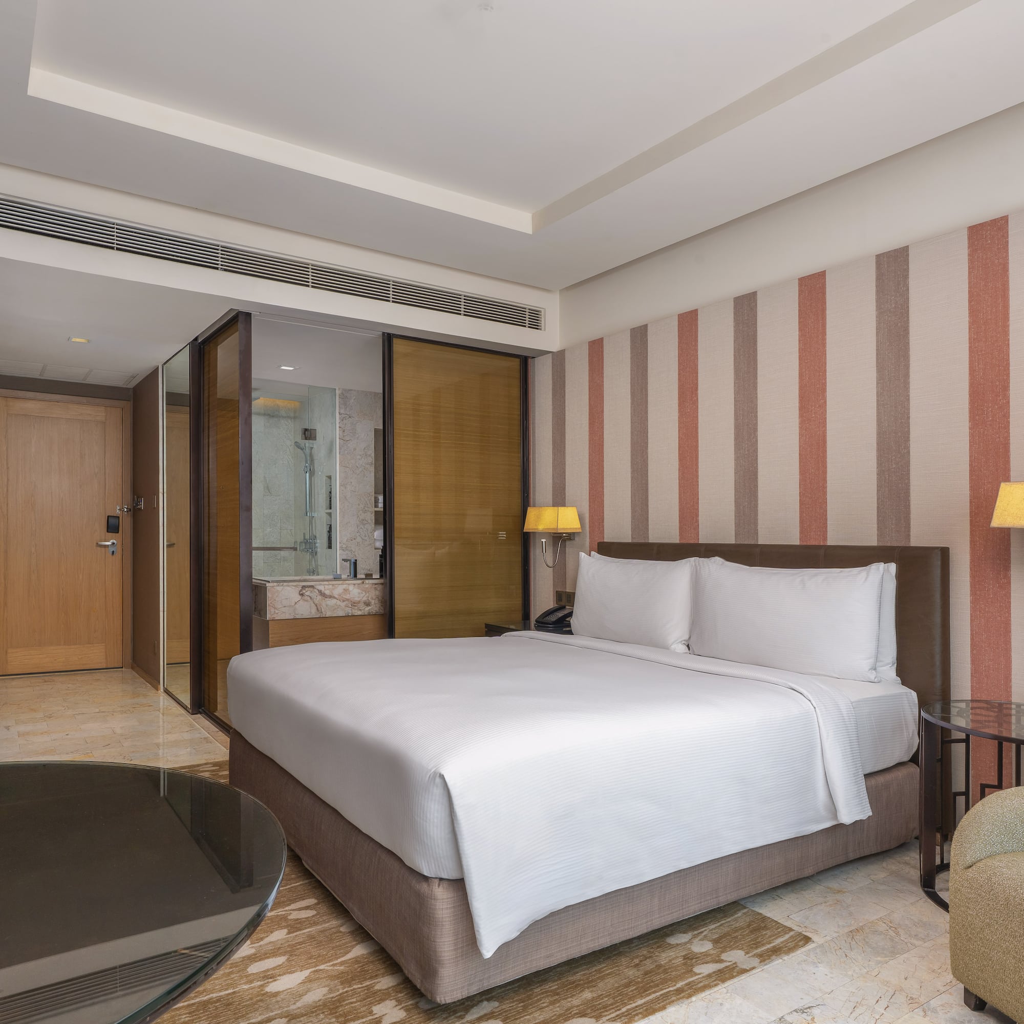 Modern hotel room with a king bed, striped accent wall, and glass-partitioned bathroom. - Guest Room - The Hotel Bangkok Sukhumvit 26 Affiliated by Meliá, Bangkok
