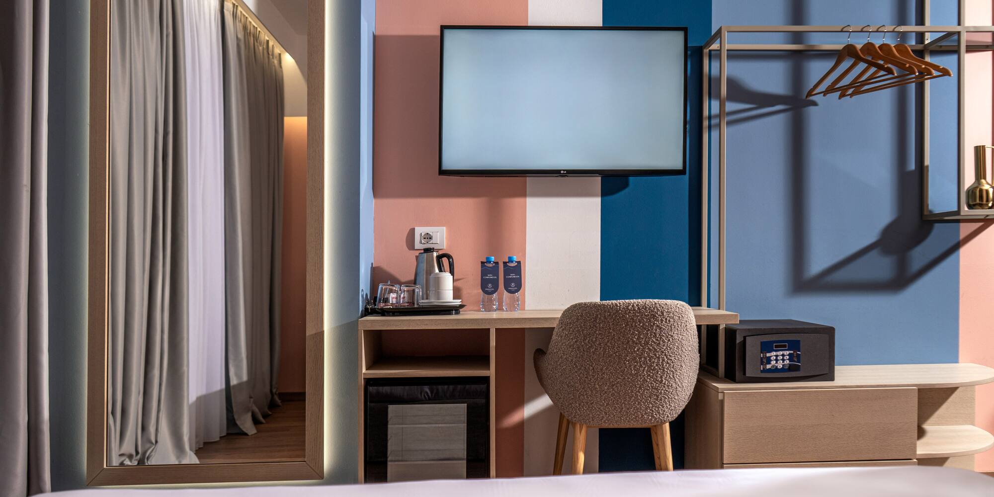 Colorful modern hotel room with TV, desk, safe, and open wardrobe.