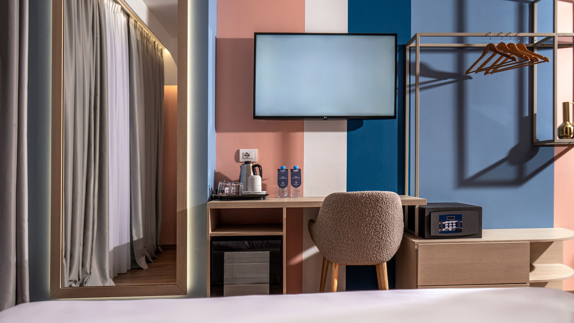 Colorful modern hotel room with TV, desk, safe, and open wardrobe.