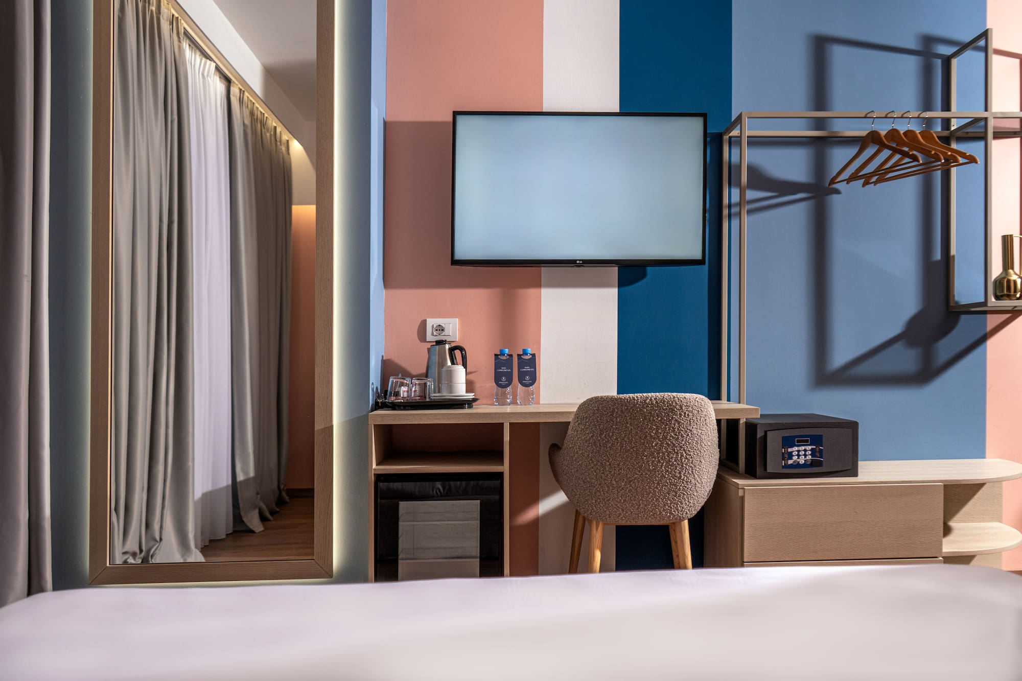 Colorful modern hotel room with TV, desk, safe, and open wardrobe.