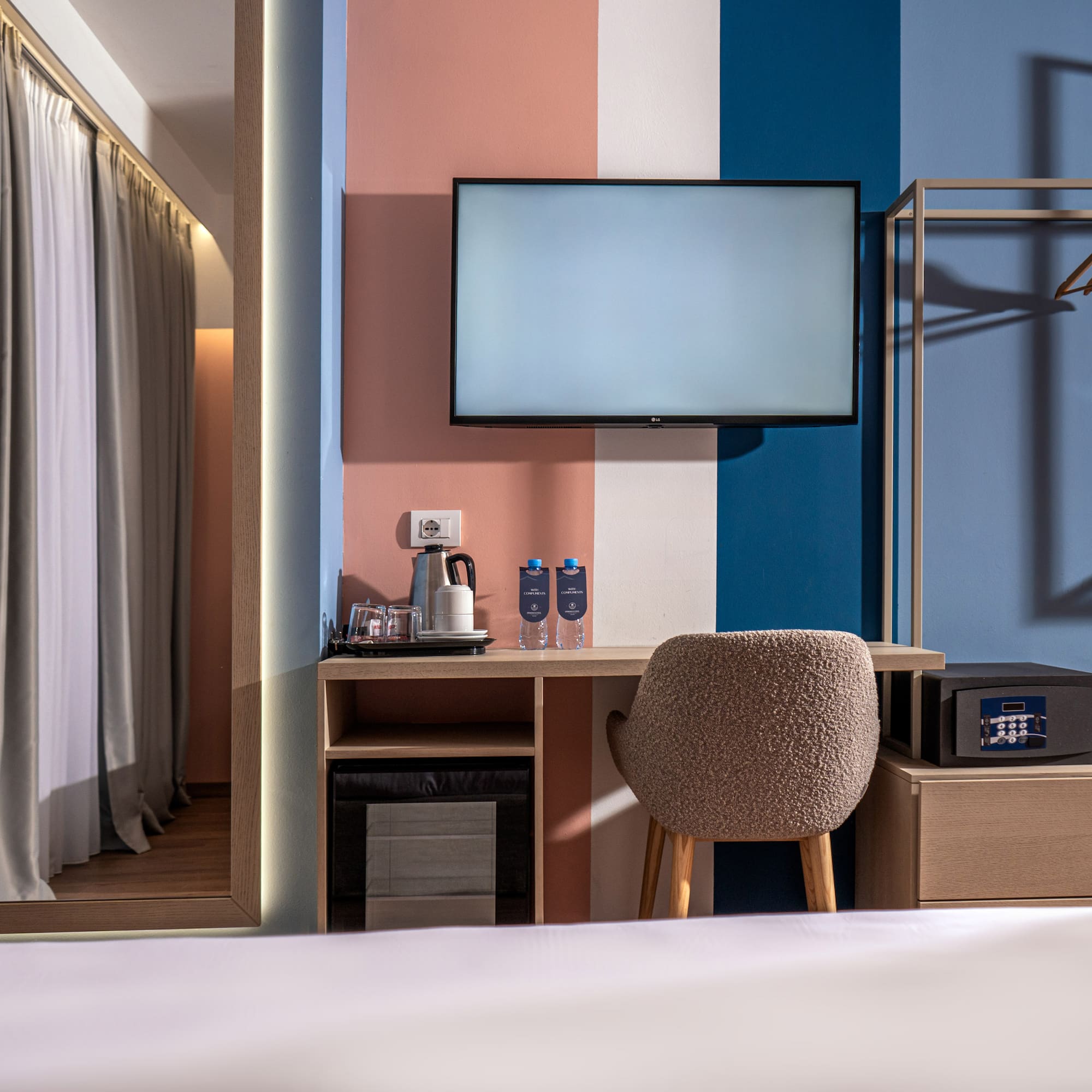 Colorful modern hotel room with TV, desk, safe, and open wardrobe.
