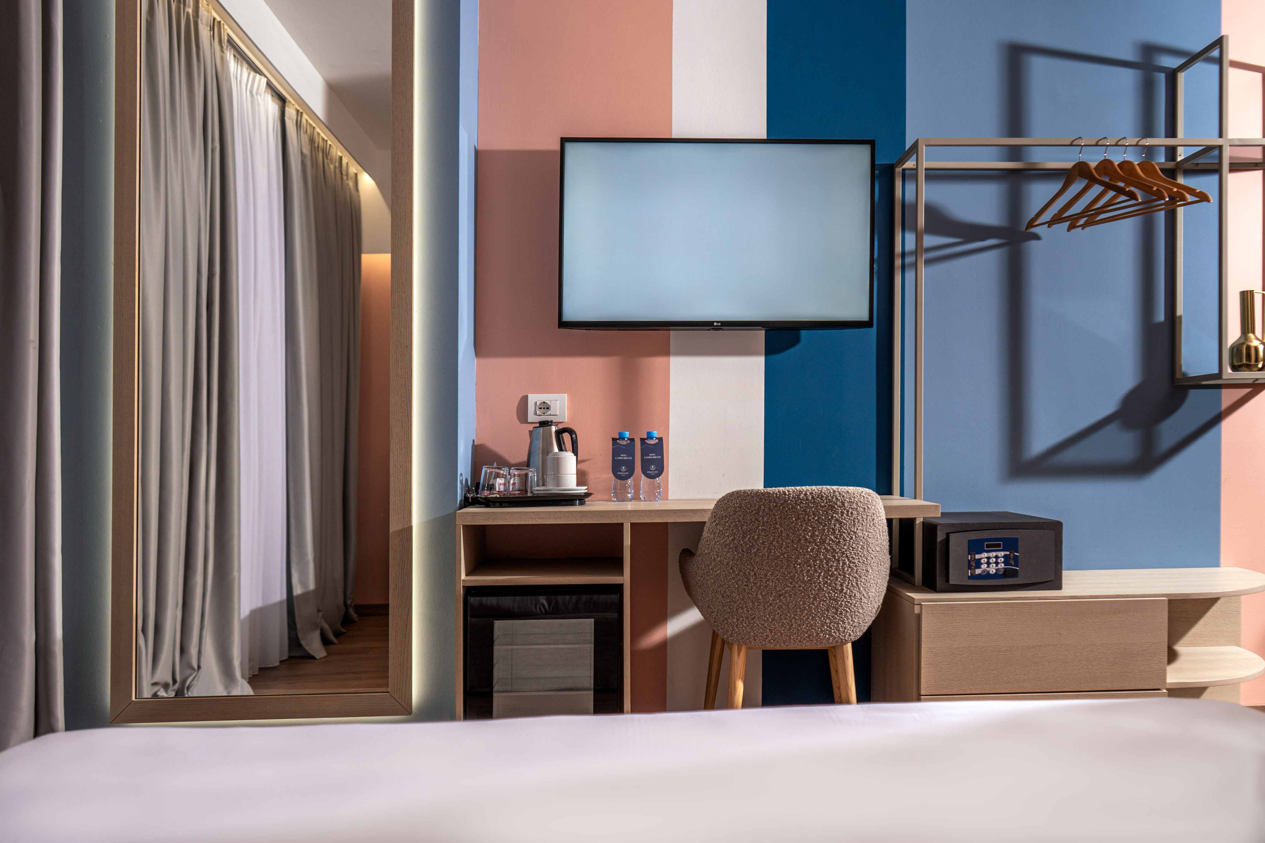 Colorful modern hotel room with TV, desk, safe, and open wardrobe.
