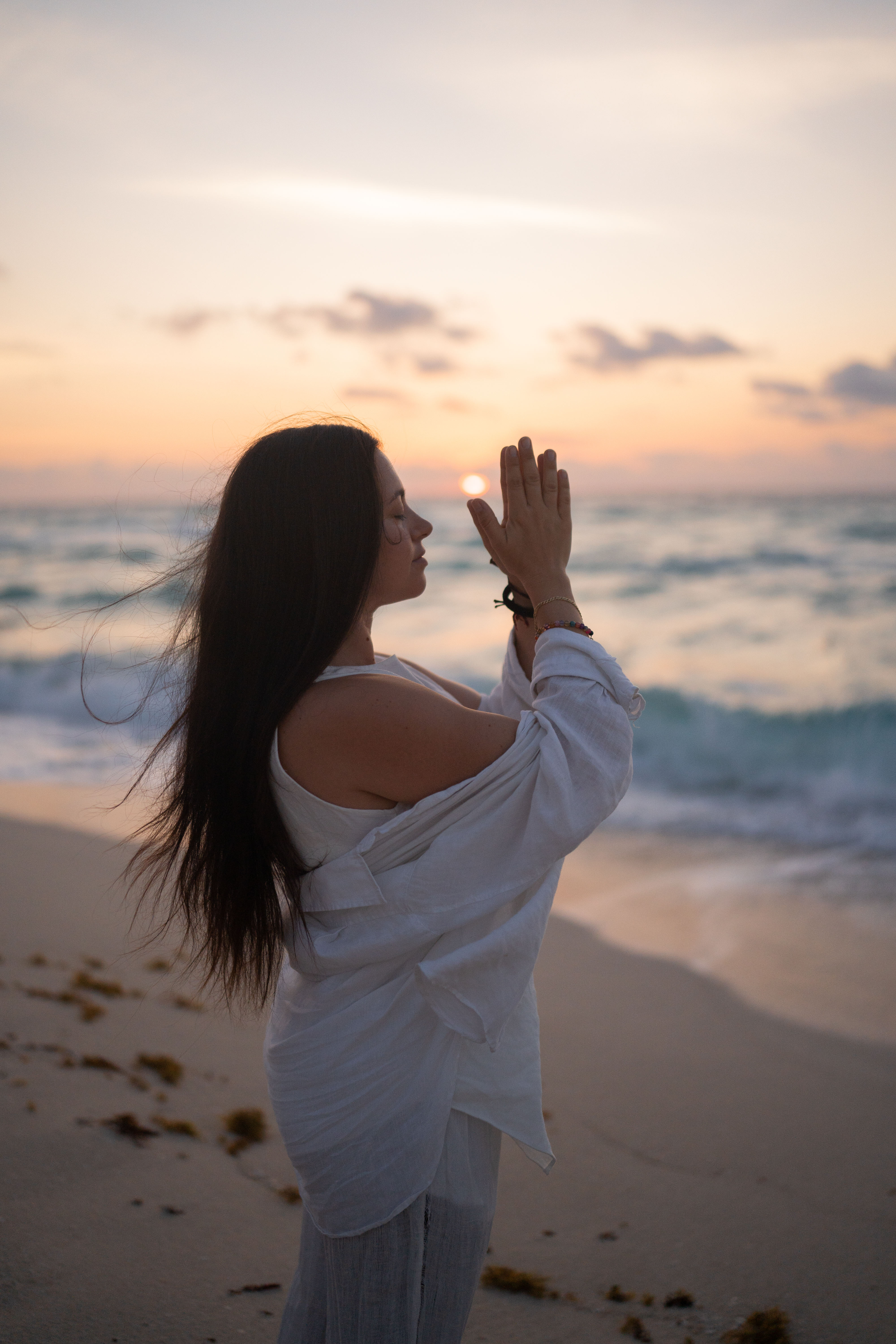 Woman meditating on beach at sunset, hands in prayer, ocean waves.