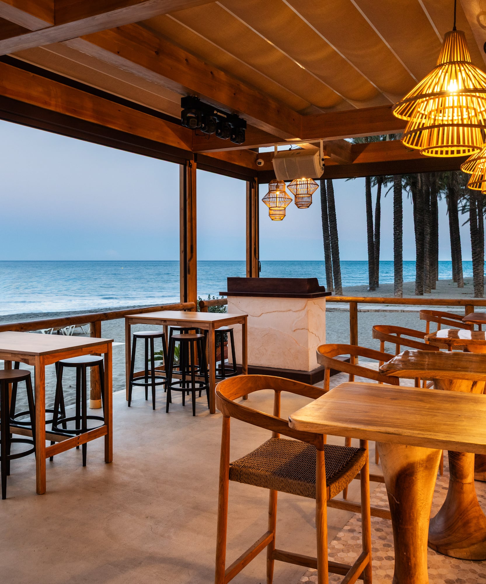 Beachfront bar with ocean view, palm trees, wooden tables, and warm lighting.