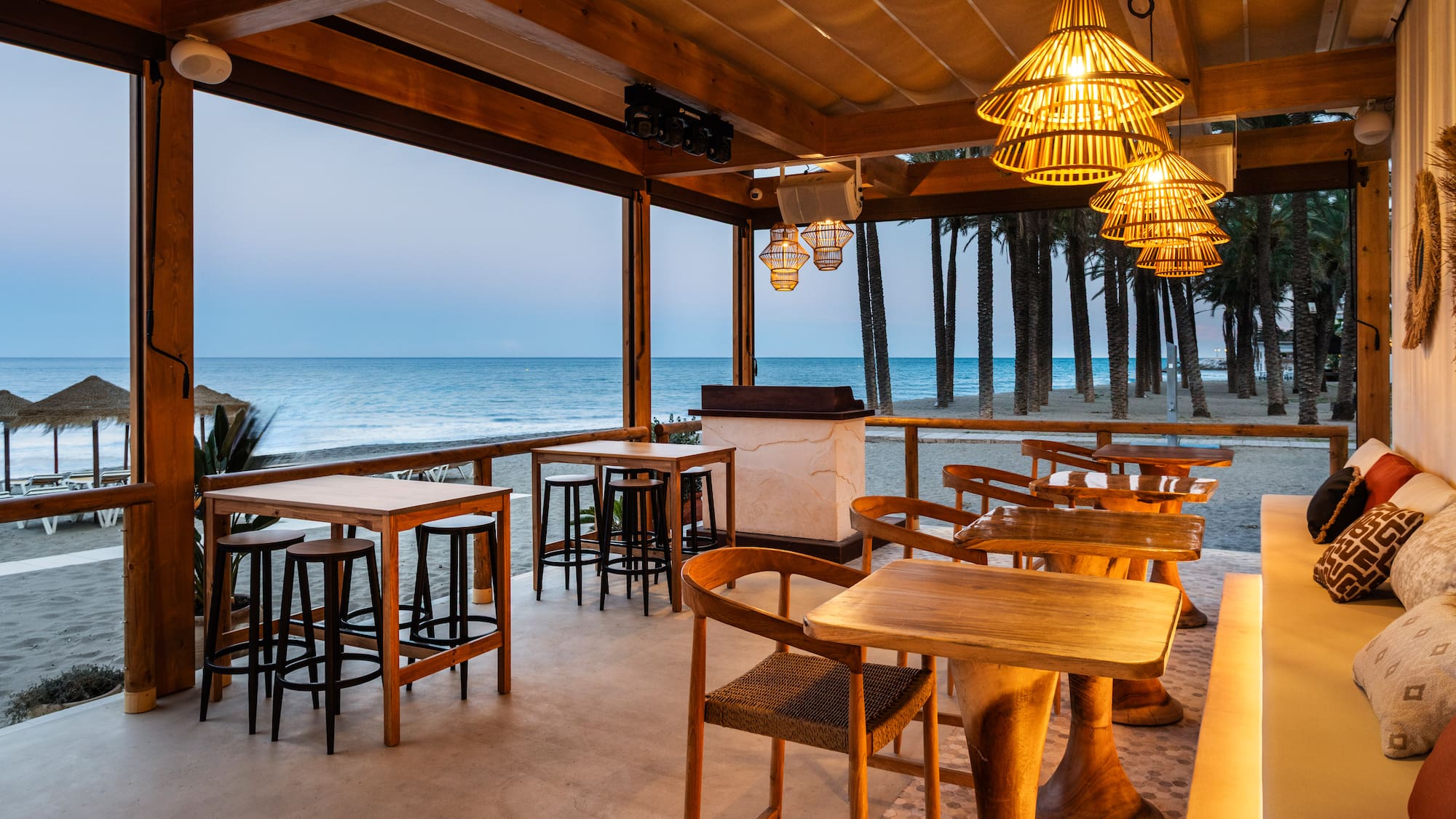 Beachfront restaurant with wooden tables, woven lights, and sea view at dusk.