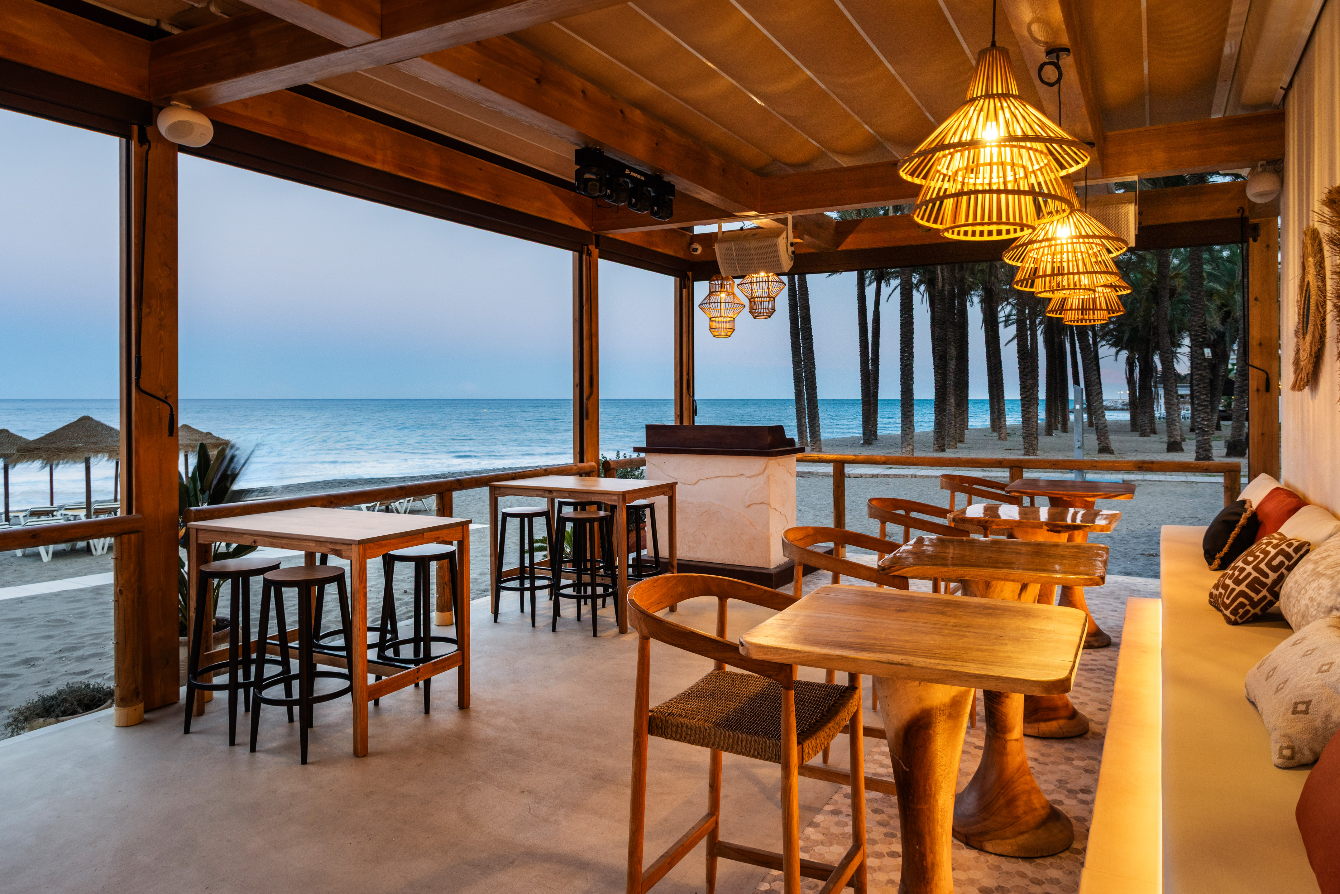 Beachfront restaurant with wooden tables, woven lights, and sea view at dusk.