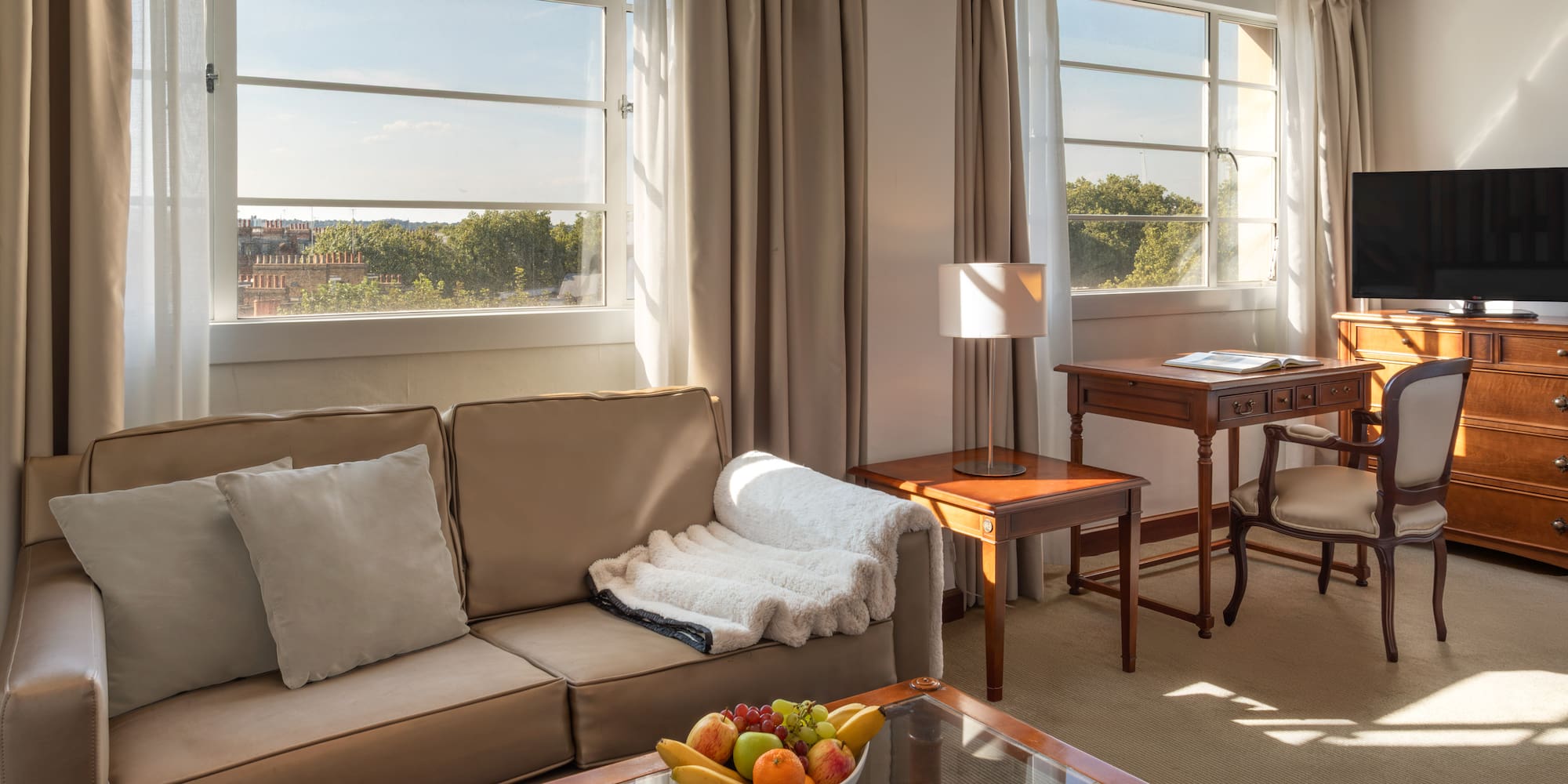 Sunlit hotel suite with cozy sofa, wooden desk, TV, and large windows.