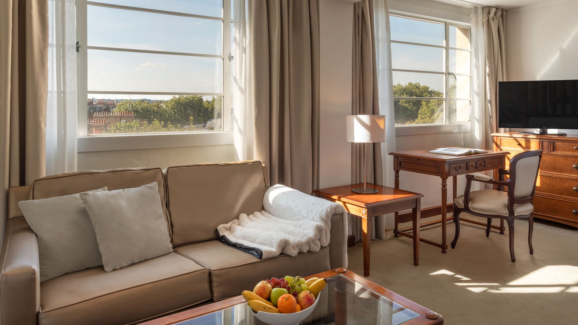 Sunlit hotel suite with cozy sofa, wooden desk, TV, and large windows.