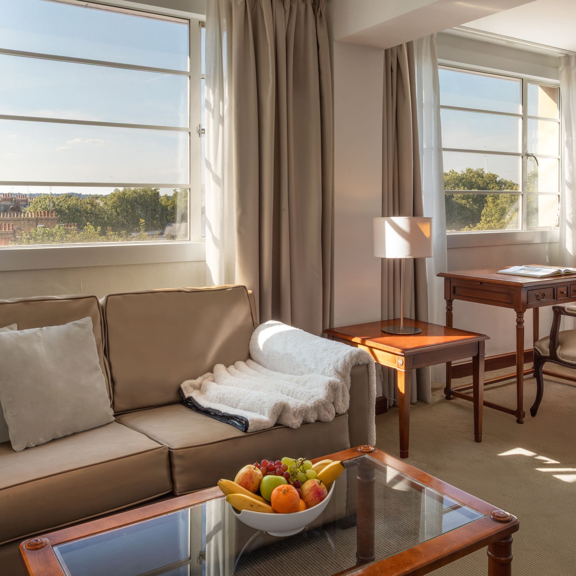 Sunlit hotel suite with cozy sofa, wooden desk, TV, and large windows.