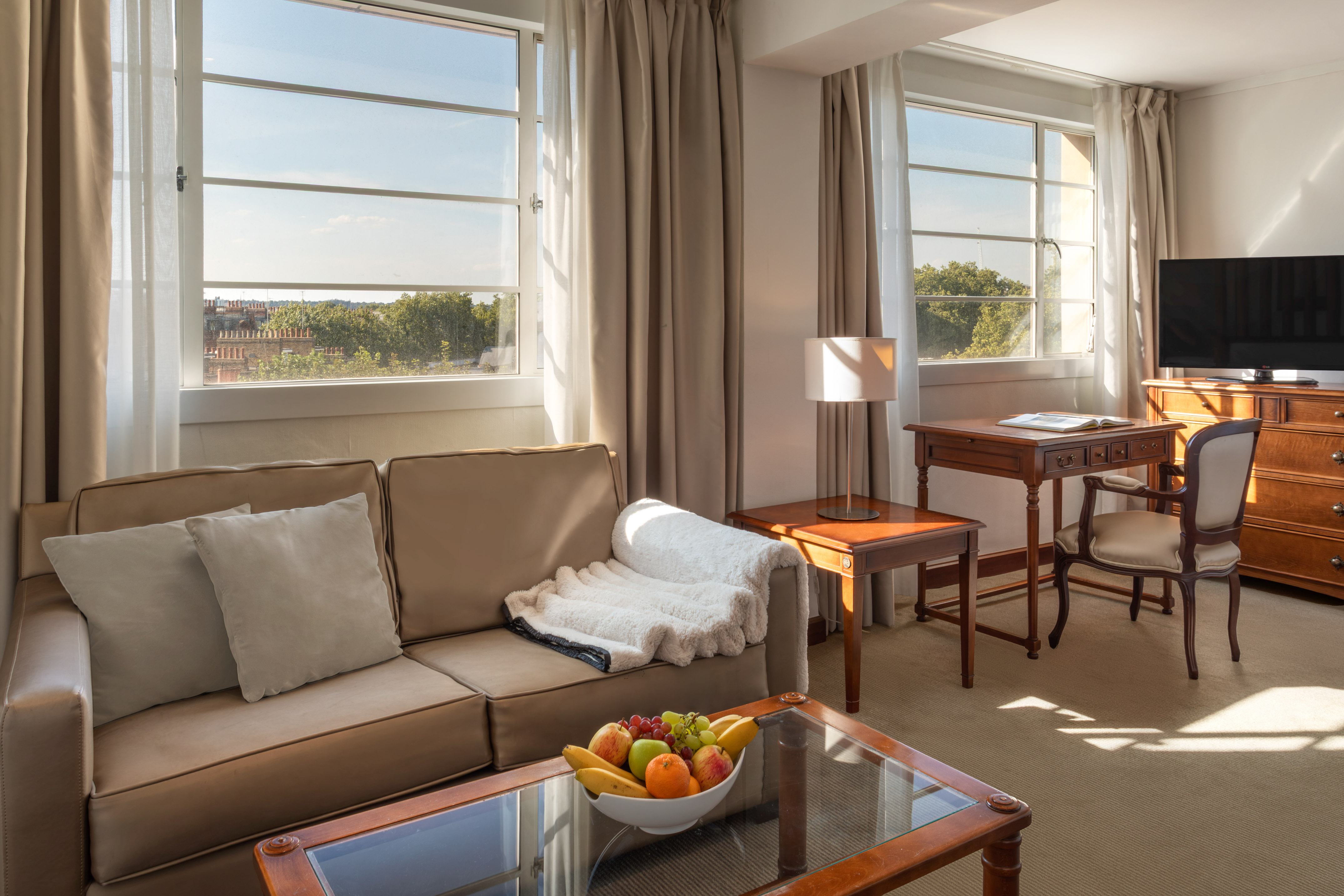 Sunlit hotel suite with cozy sofa, wooden desk, TV, and large windows.