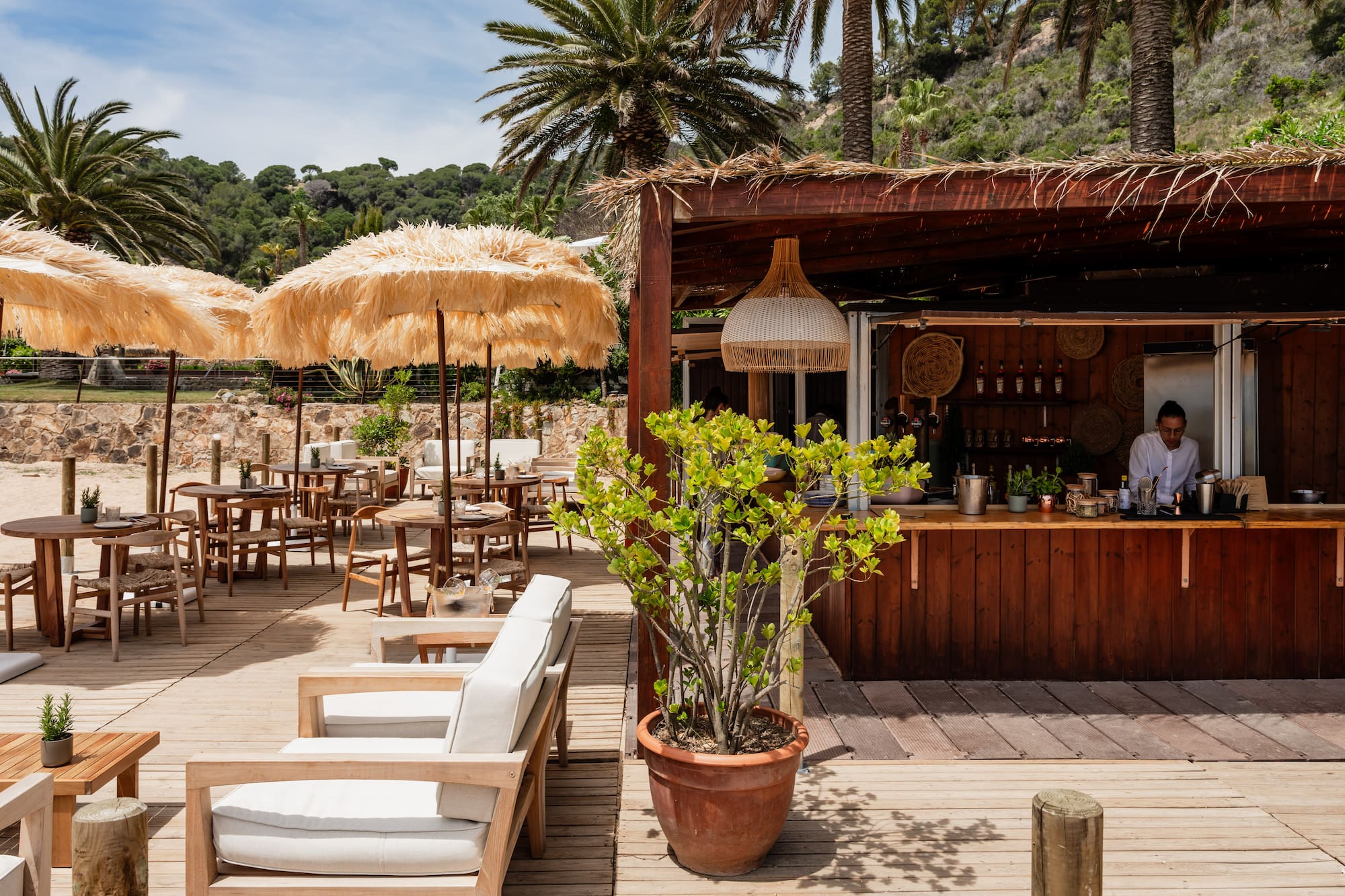 Beach bar deck with straw umbrellas, outdoor seating, and palm trees.