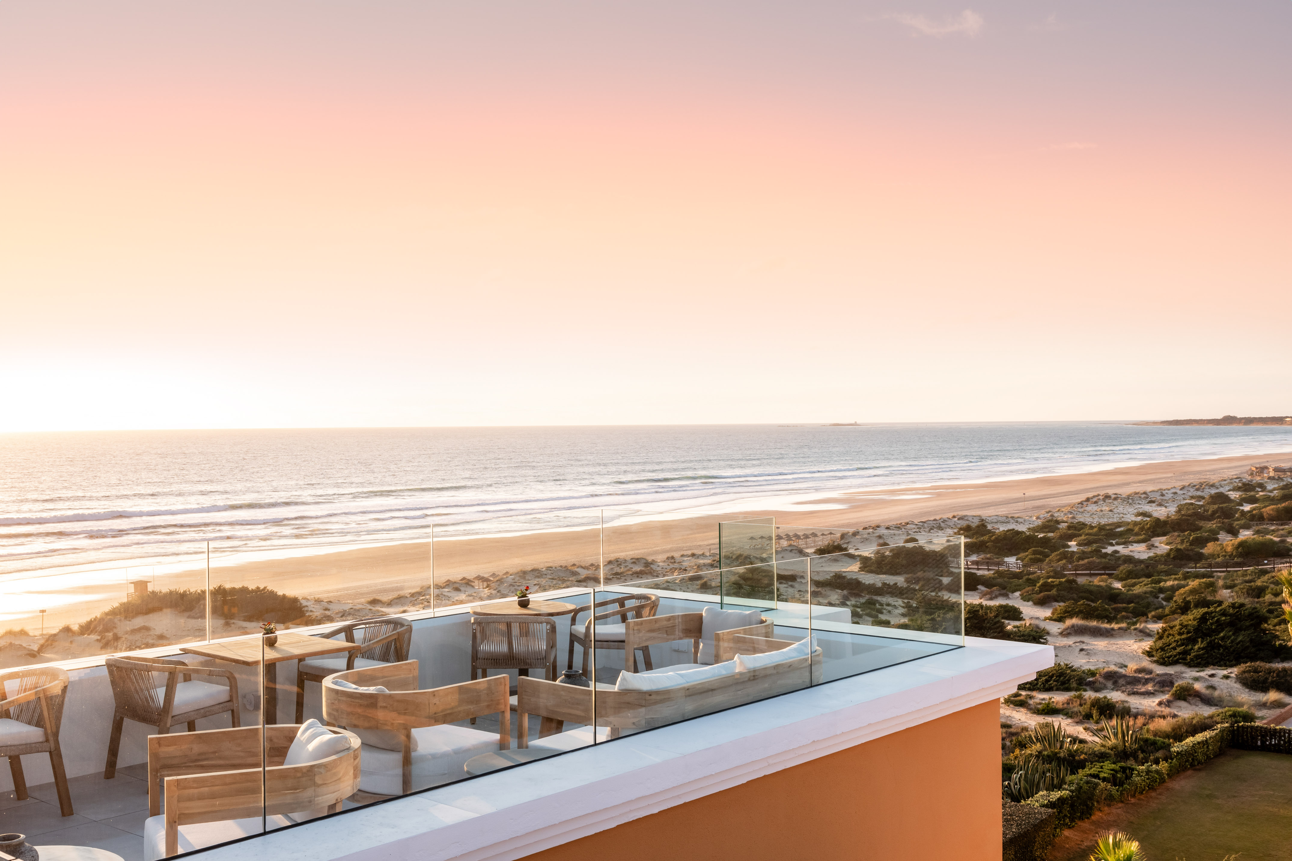 Coastal rooftop terrace with outdoor seating, ocean, beach, and sunset.