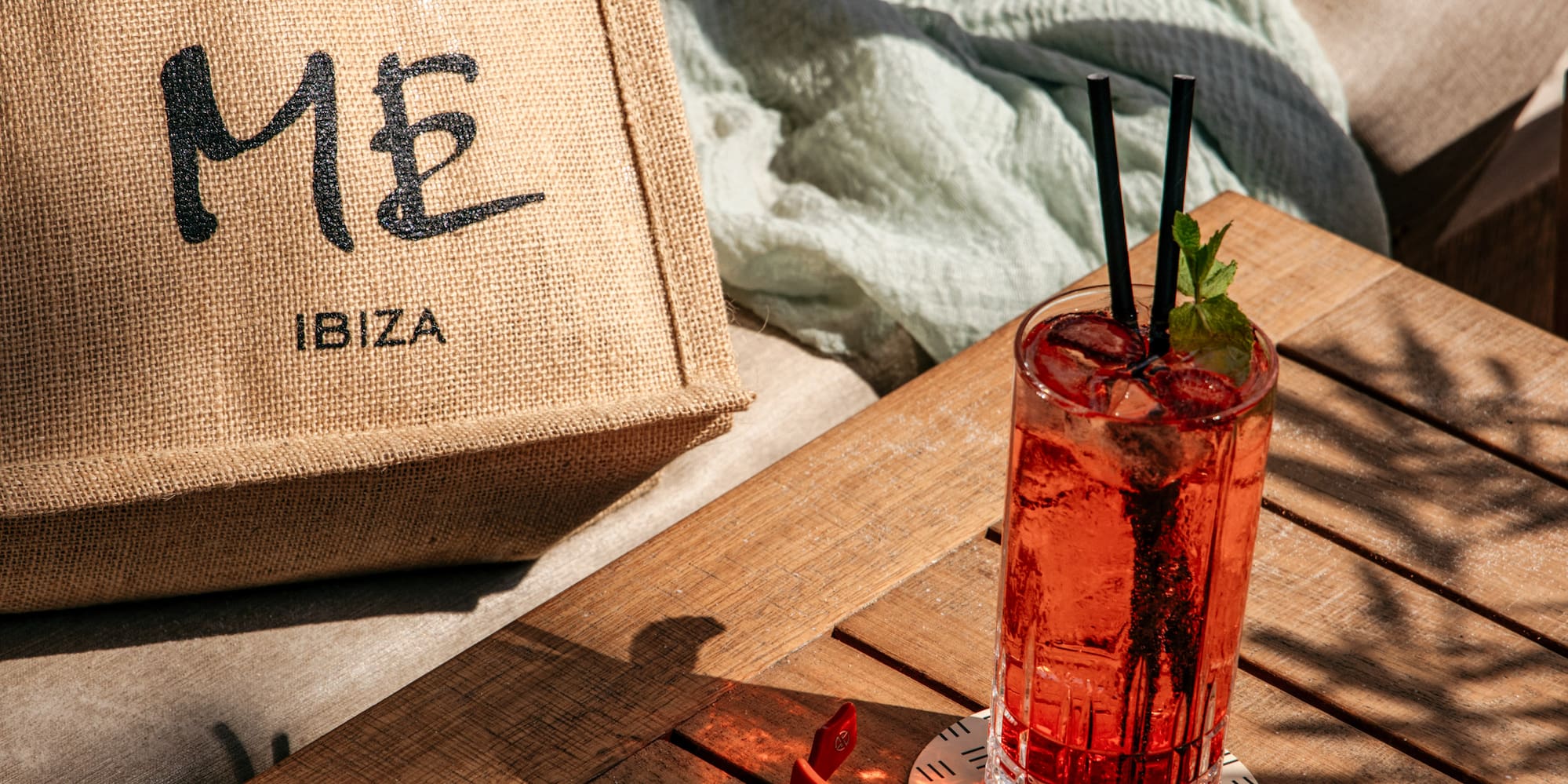 Red cocktail, orange sunglasses, and ME Ibiza tote bag on a sunlit wooden table.