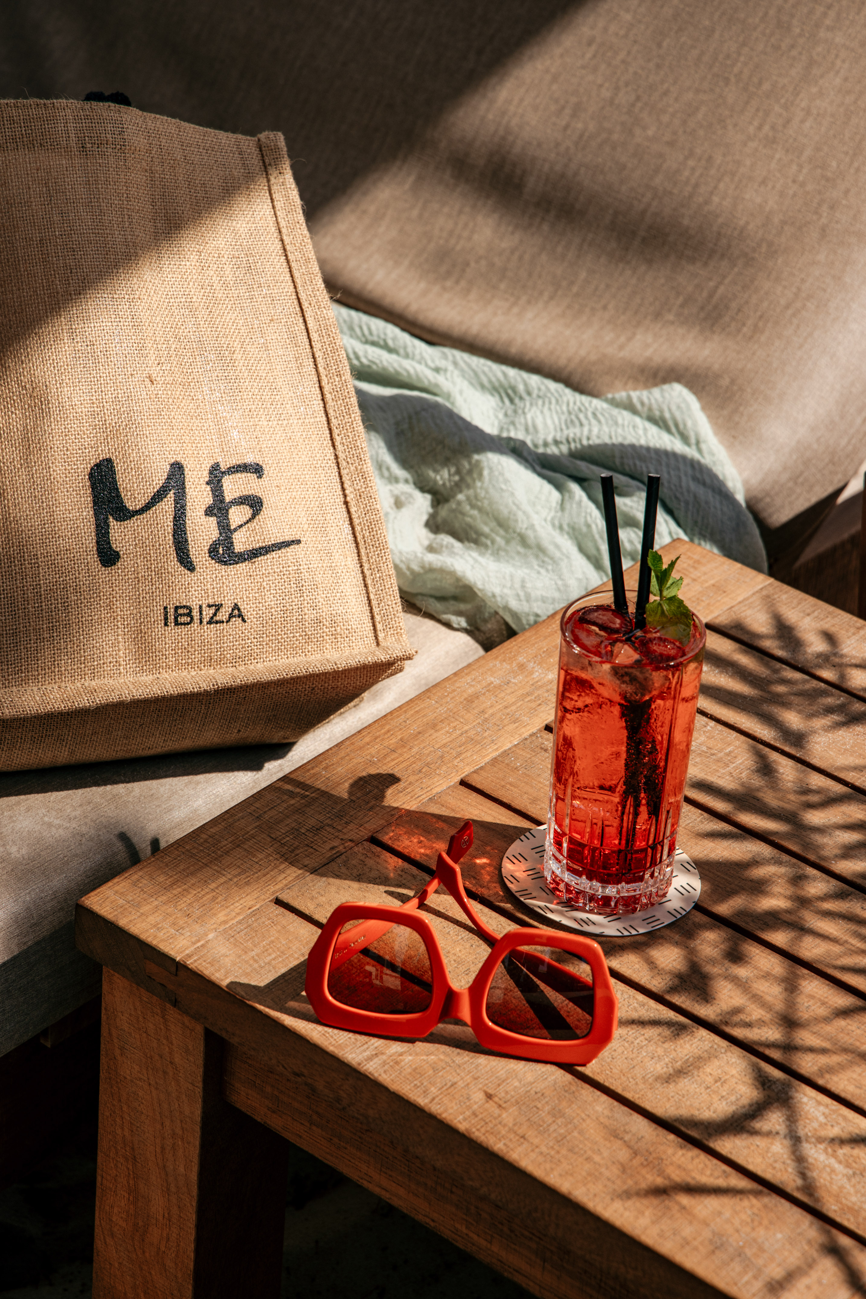 Red cocktail, orange sunglasses, and ME Ibiza tote bag on a sunlit wooden table.