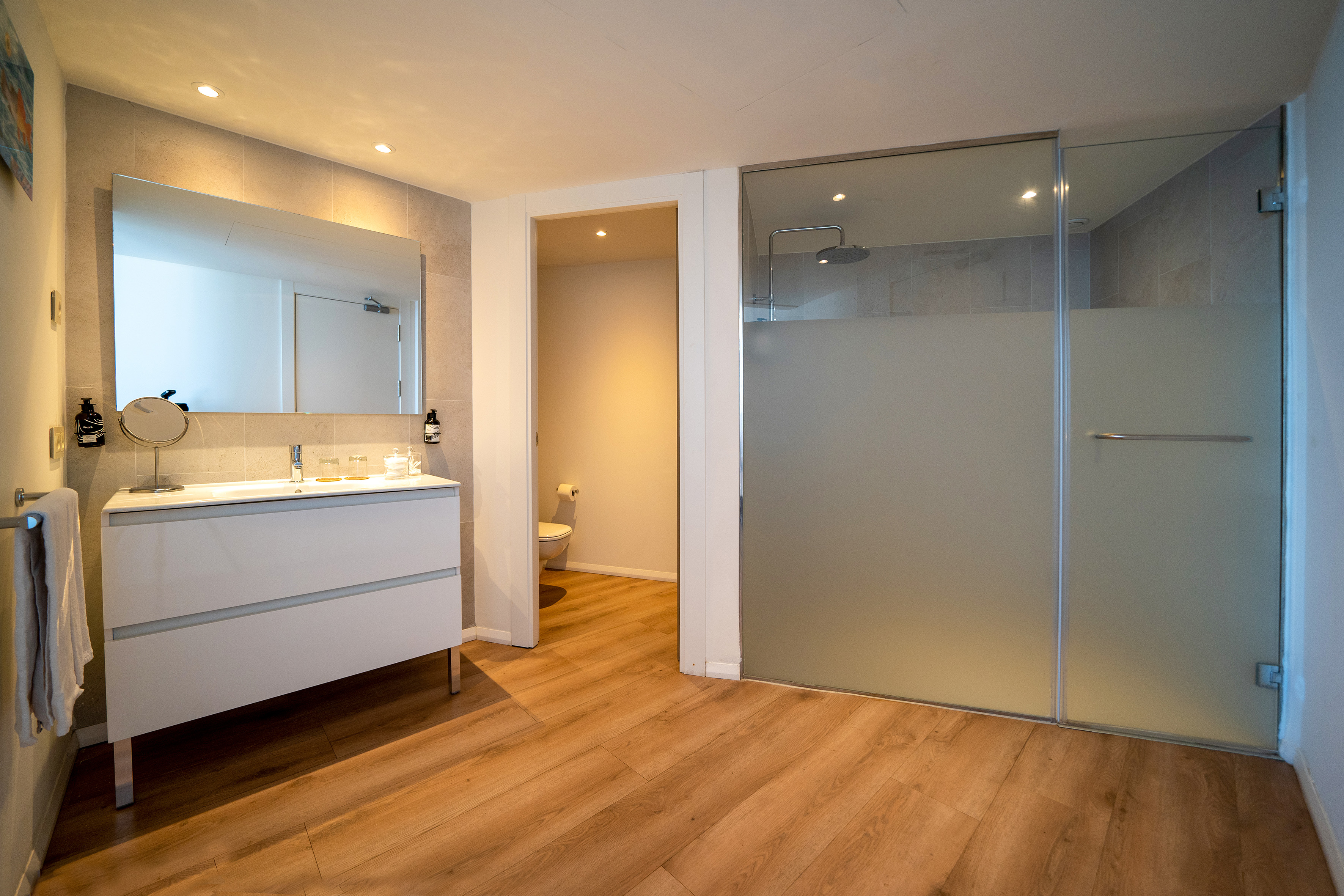 Modern bathroom with white vanity, large mirror, wood floor, glass shower.