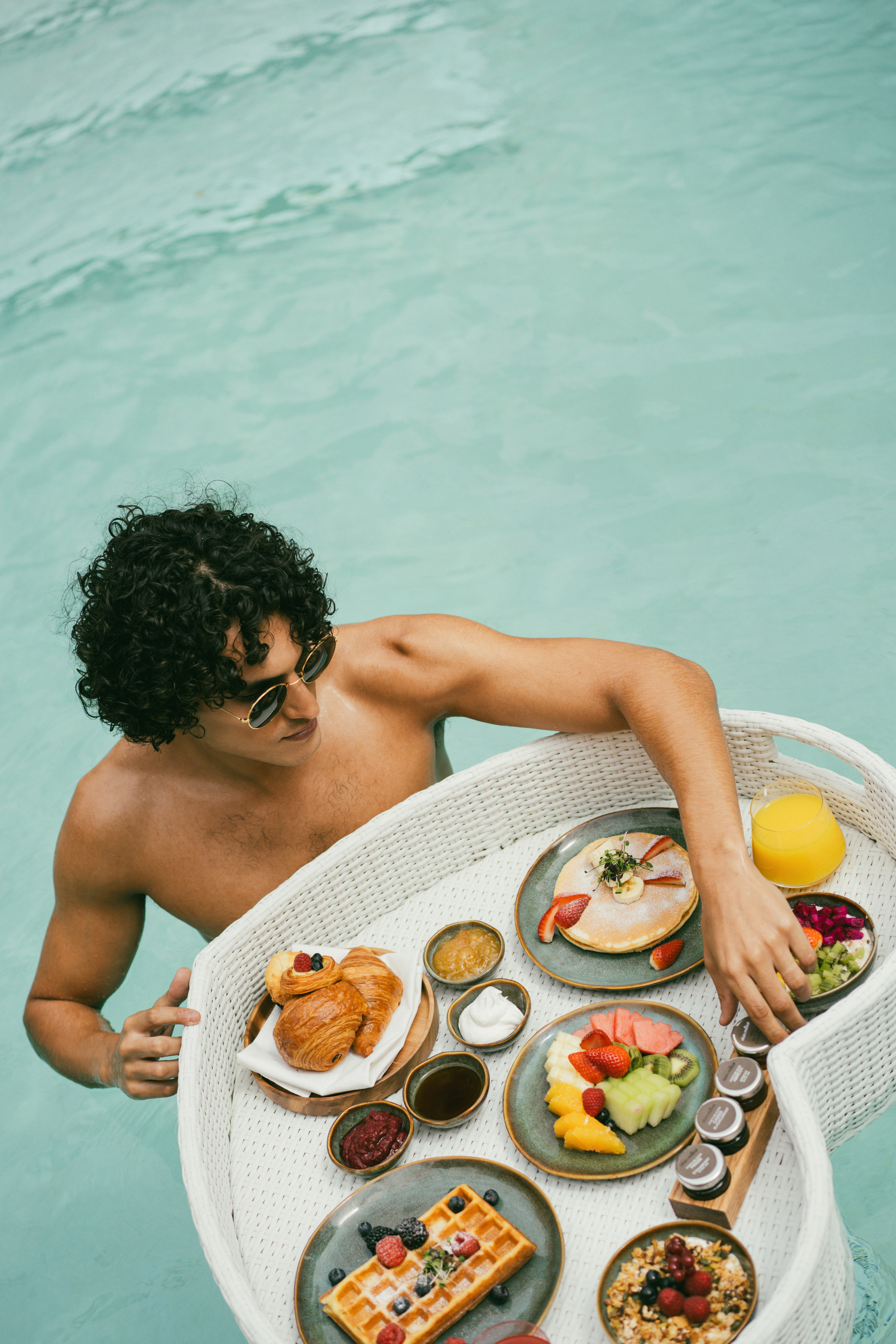 Man in pool enjoys floating breakfast: pancakes, waffles, fruit, croissants.