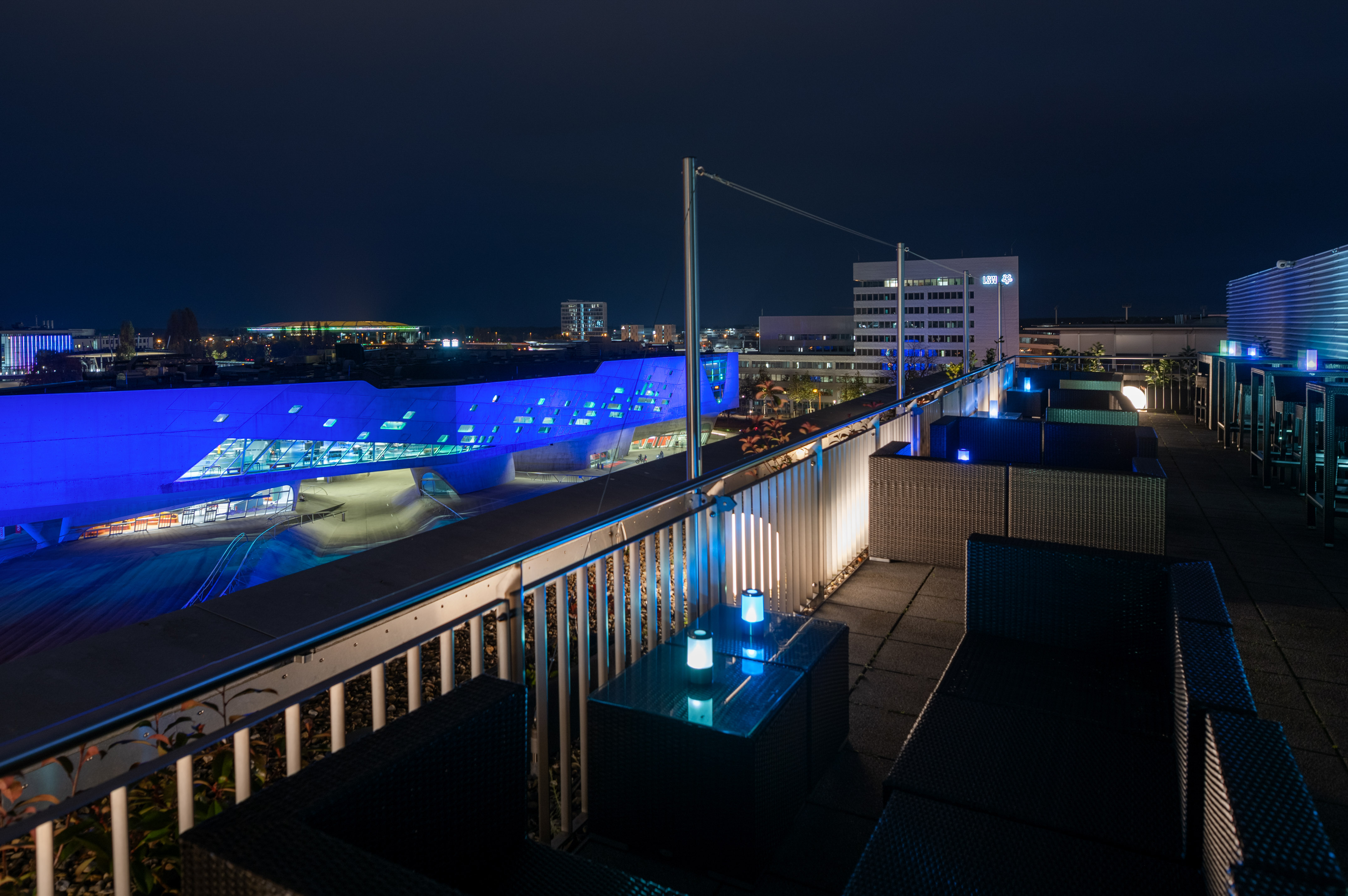 Nighttime rooftop lounge with outdoor seating overlooking blue illuminated city. - Sky Lounge & Bar The VIEW, Aperitivos - INNSiDE Wolfsburg, Wolfsburg