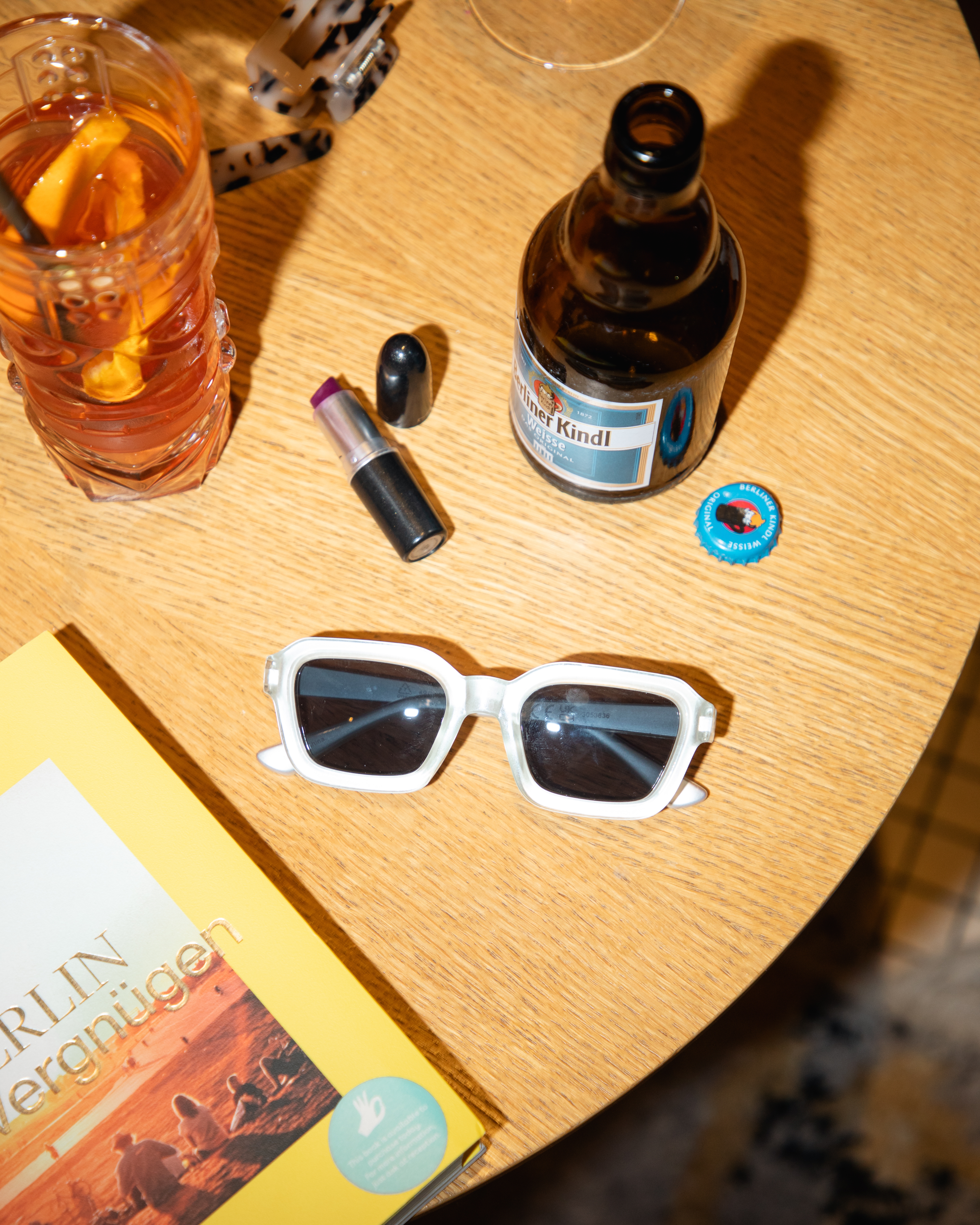 White sunglasses, cocktail, Berliner Kindl beer, lipstick, and Berlin book on table.