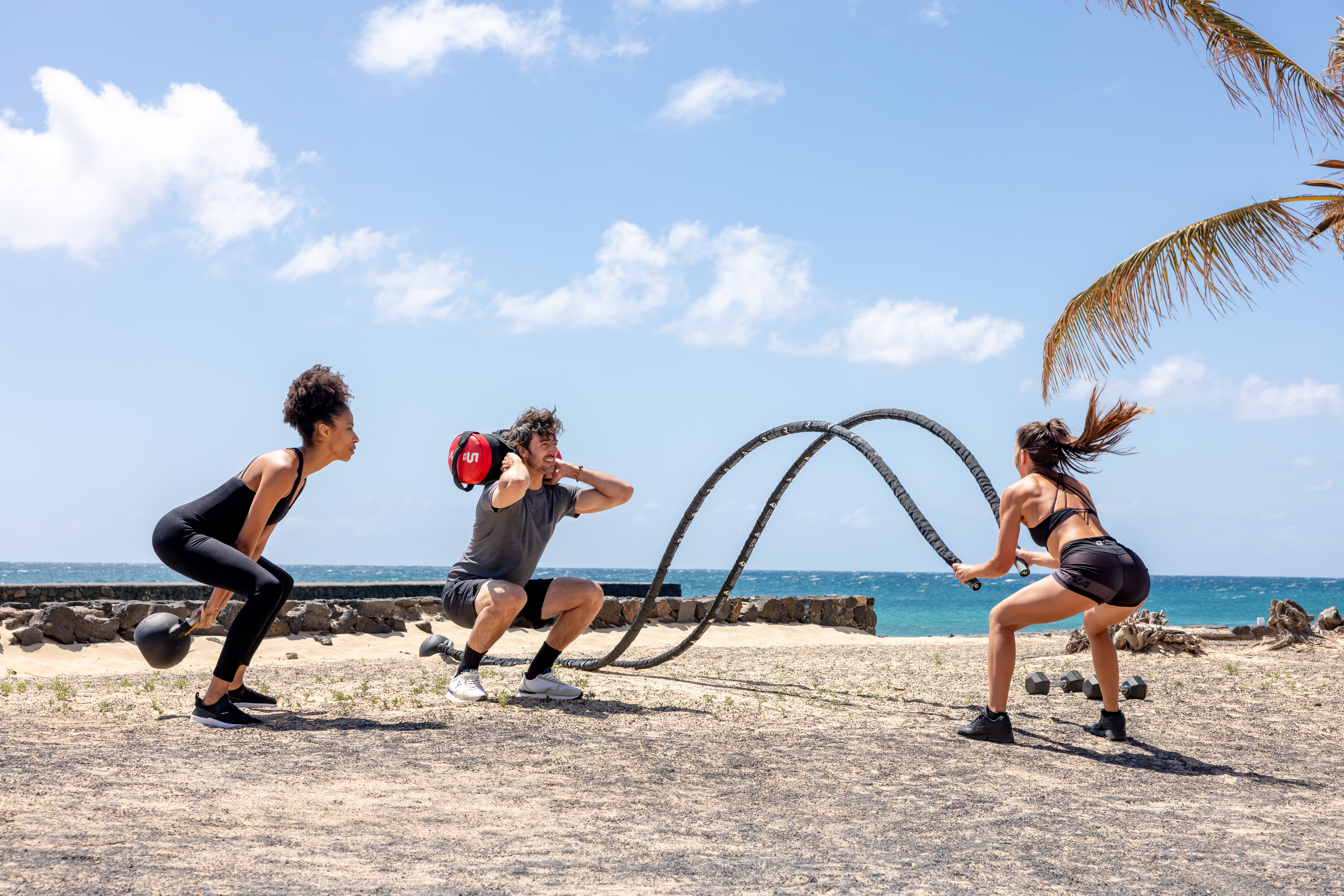 Outdoor beach fitness: three people with kettlebell, sandbag, battle ropes.