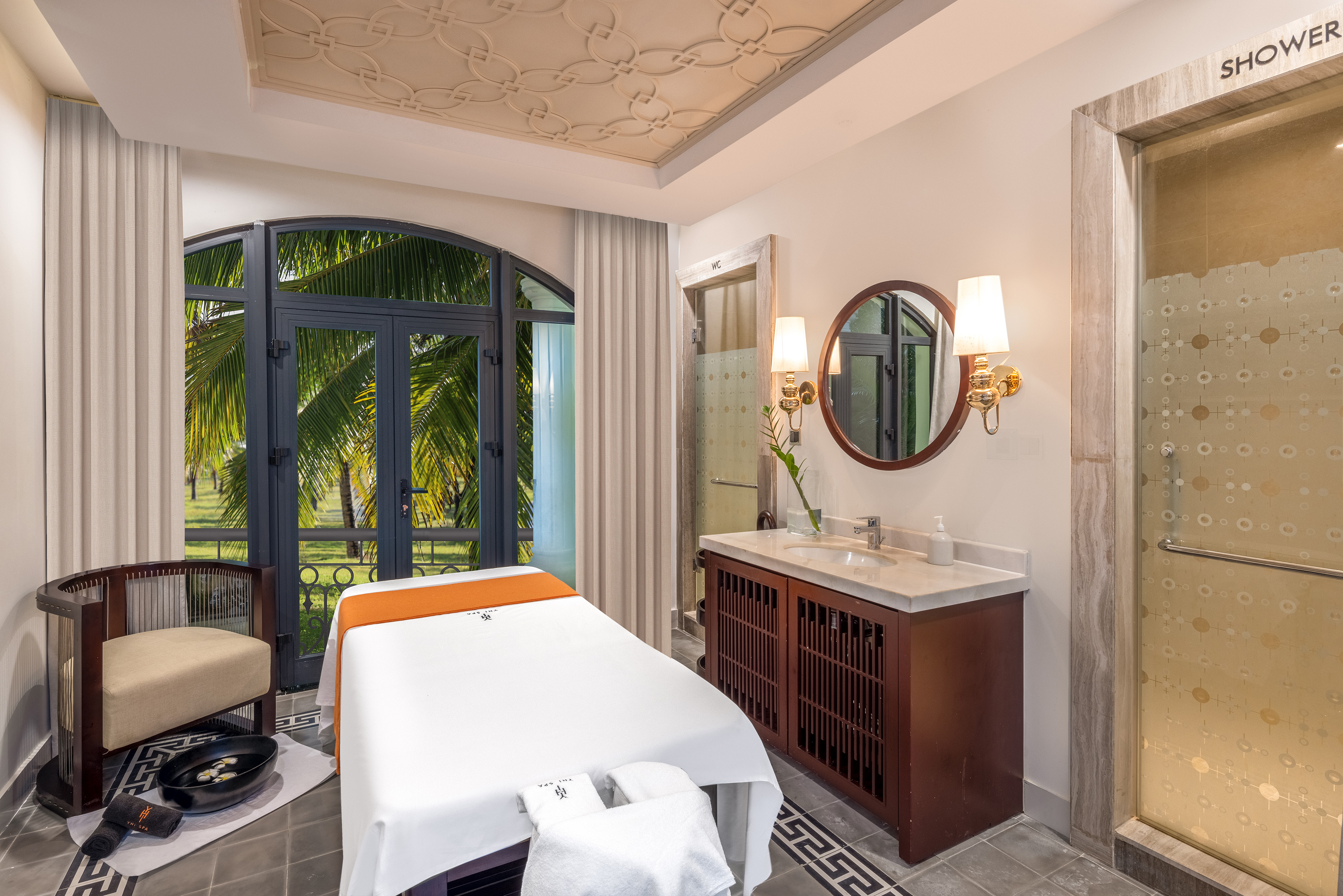 Spa treatment room with massage bed, vanity, shower, and tropical palm view.