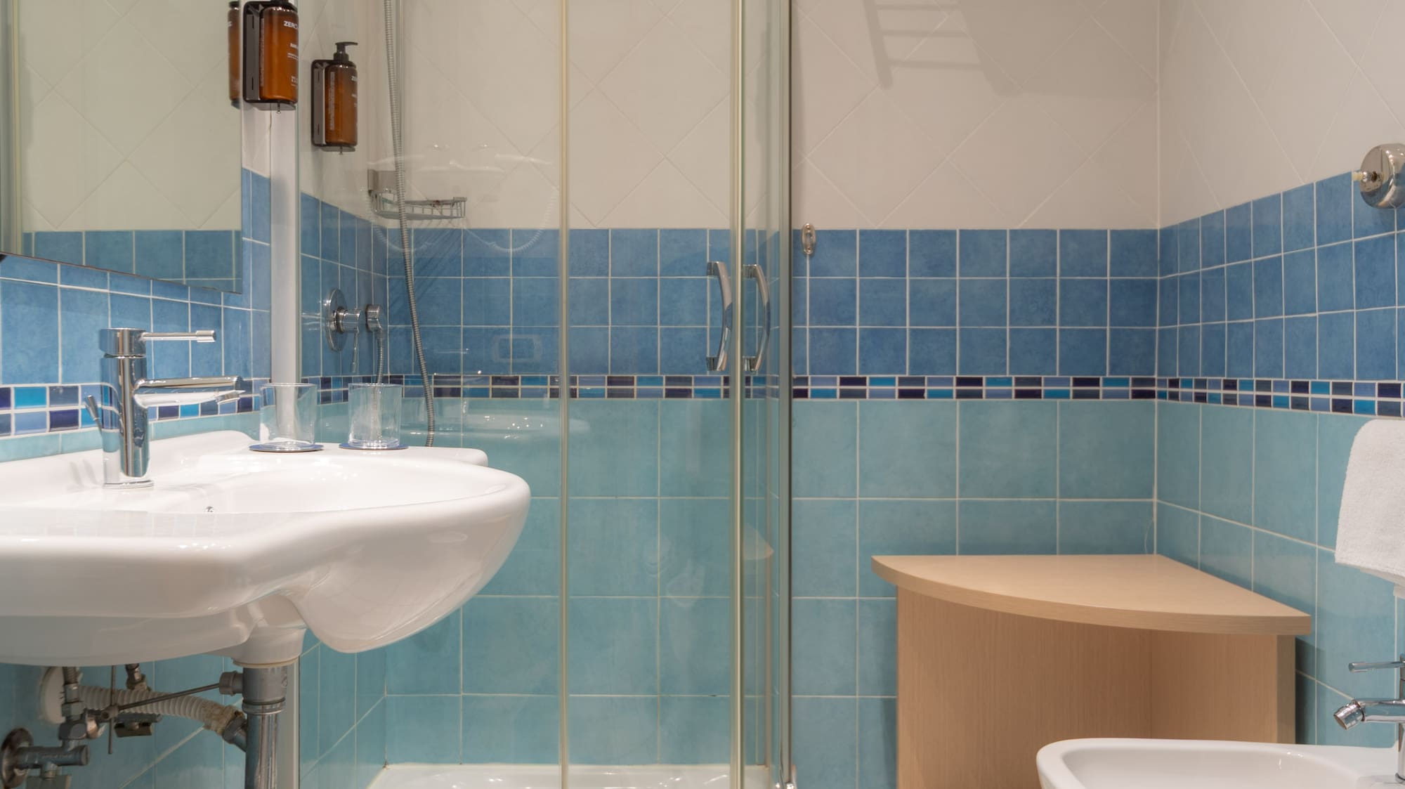 Blue and white tiled bathroom with glass shower, sink, toilet, and bidet.