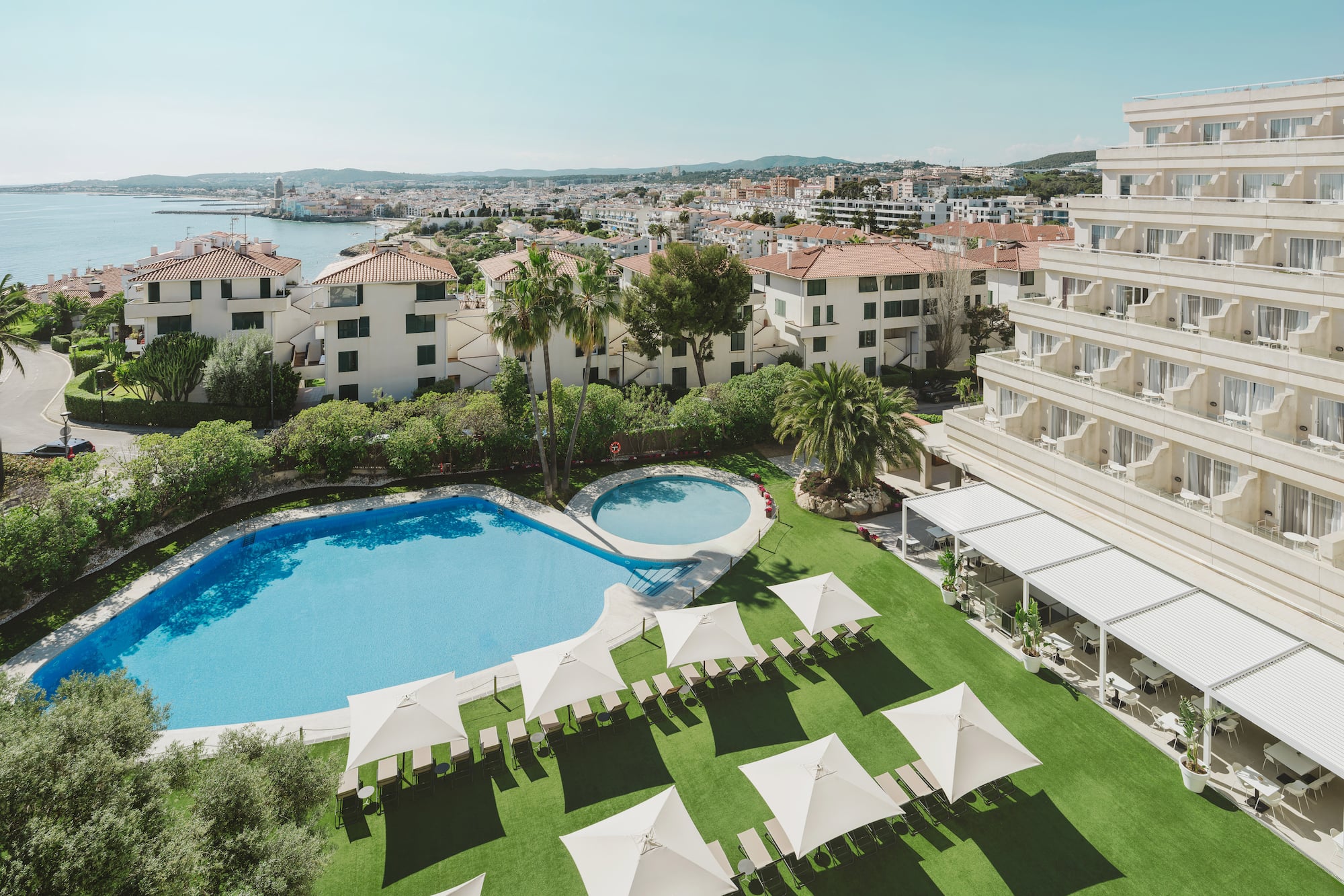Resort con piscina, palmeras y vistas al mar y ciudad costera.