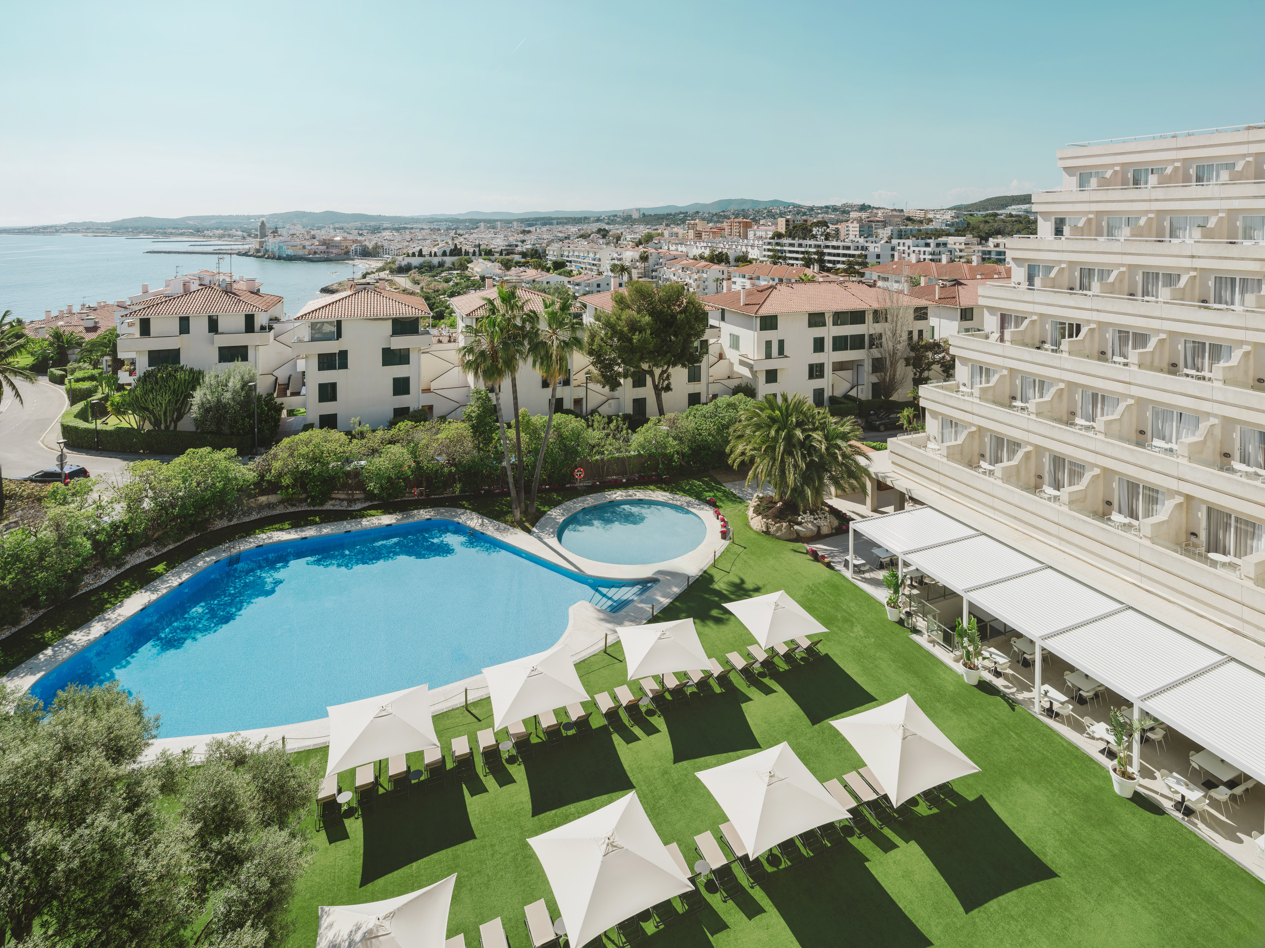 Resort con piscina, palmeras y vistas al mar y ciudad costera.