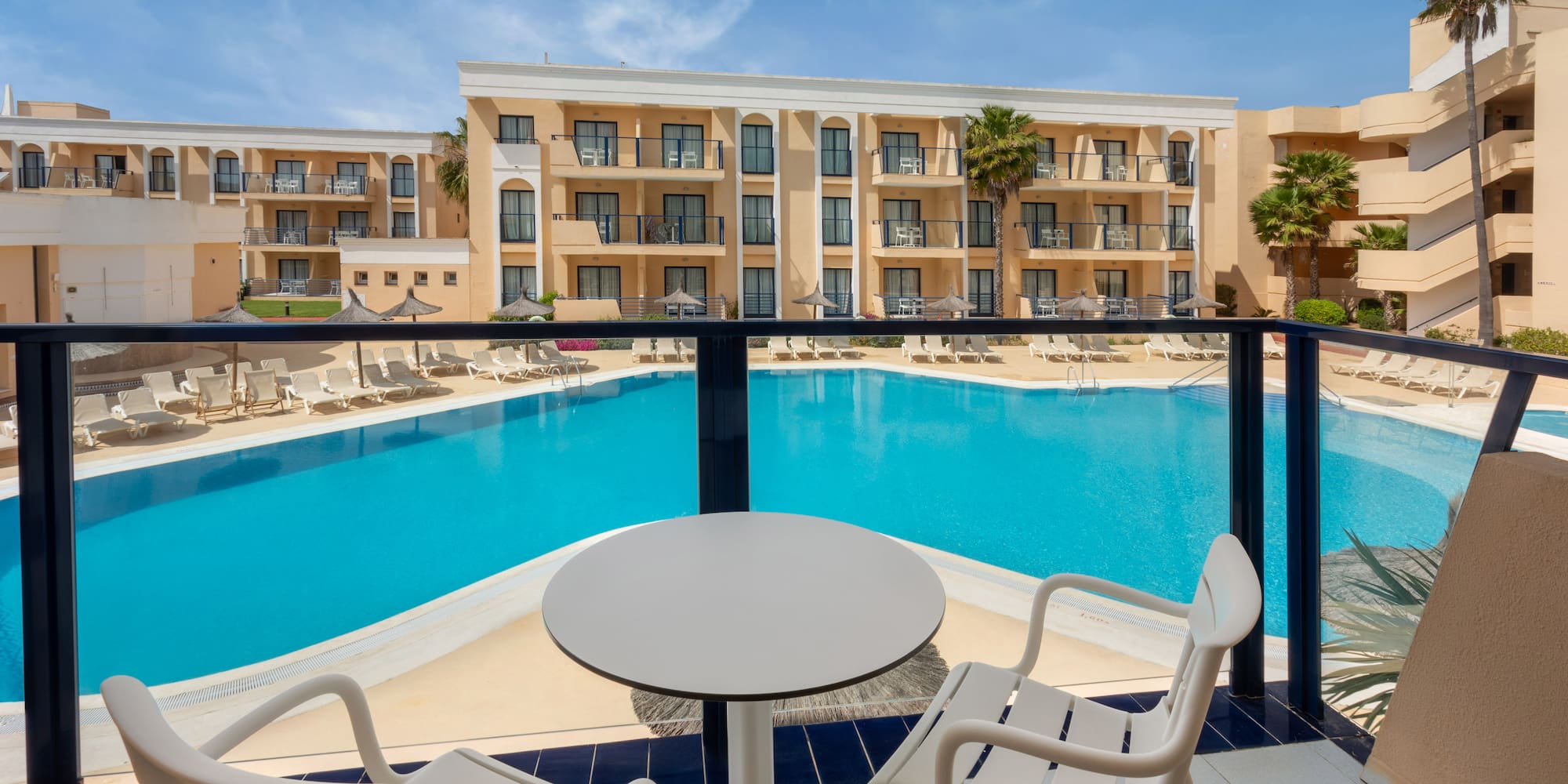 Resort hotel balcony view: large swimming pool, palm trees, sun loungers.