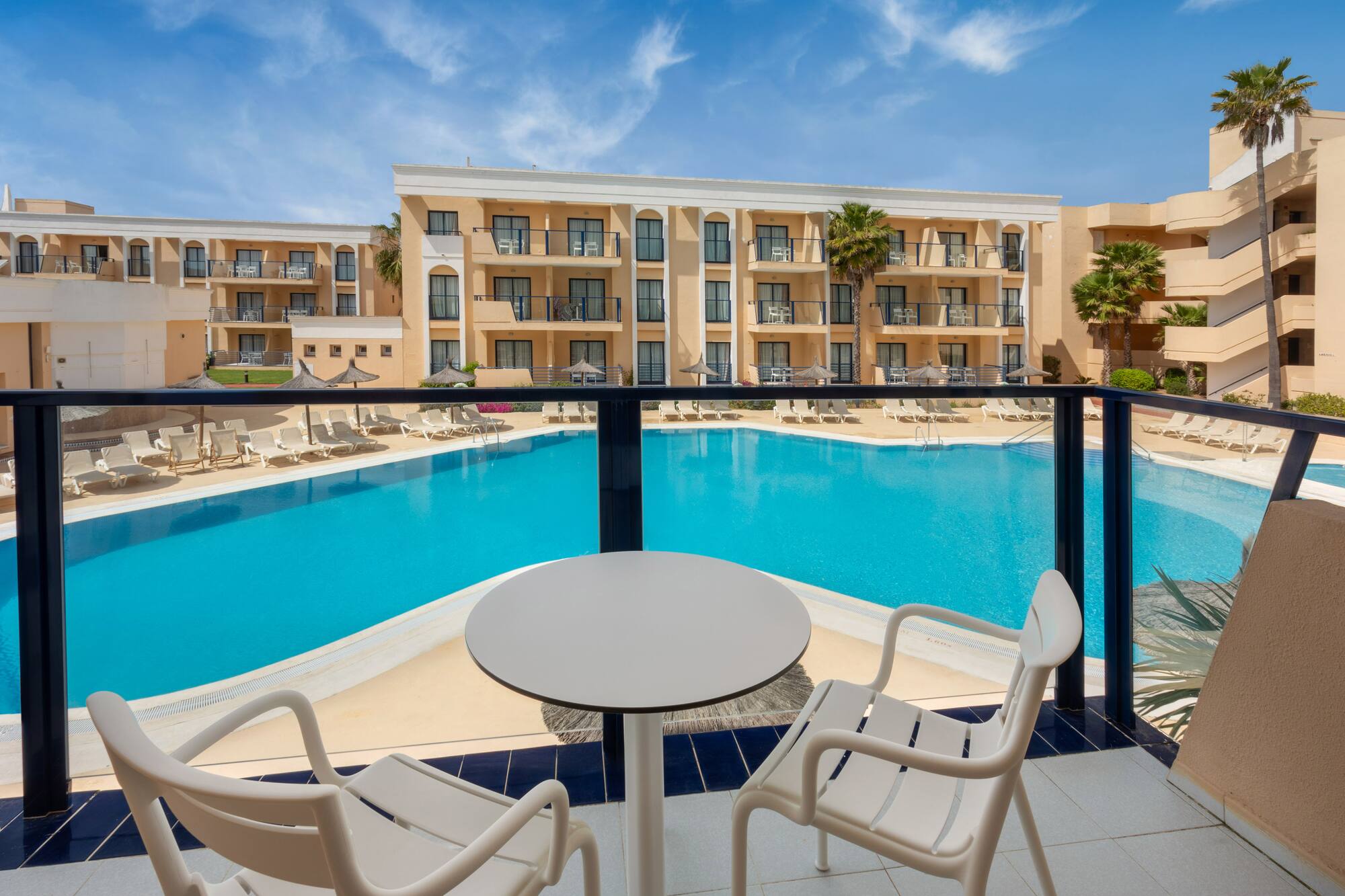 Resort hotel balcony view: large swimming pool, palm trees, sun loungers.