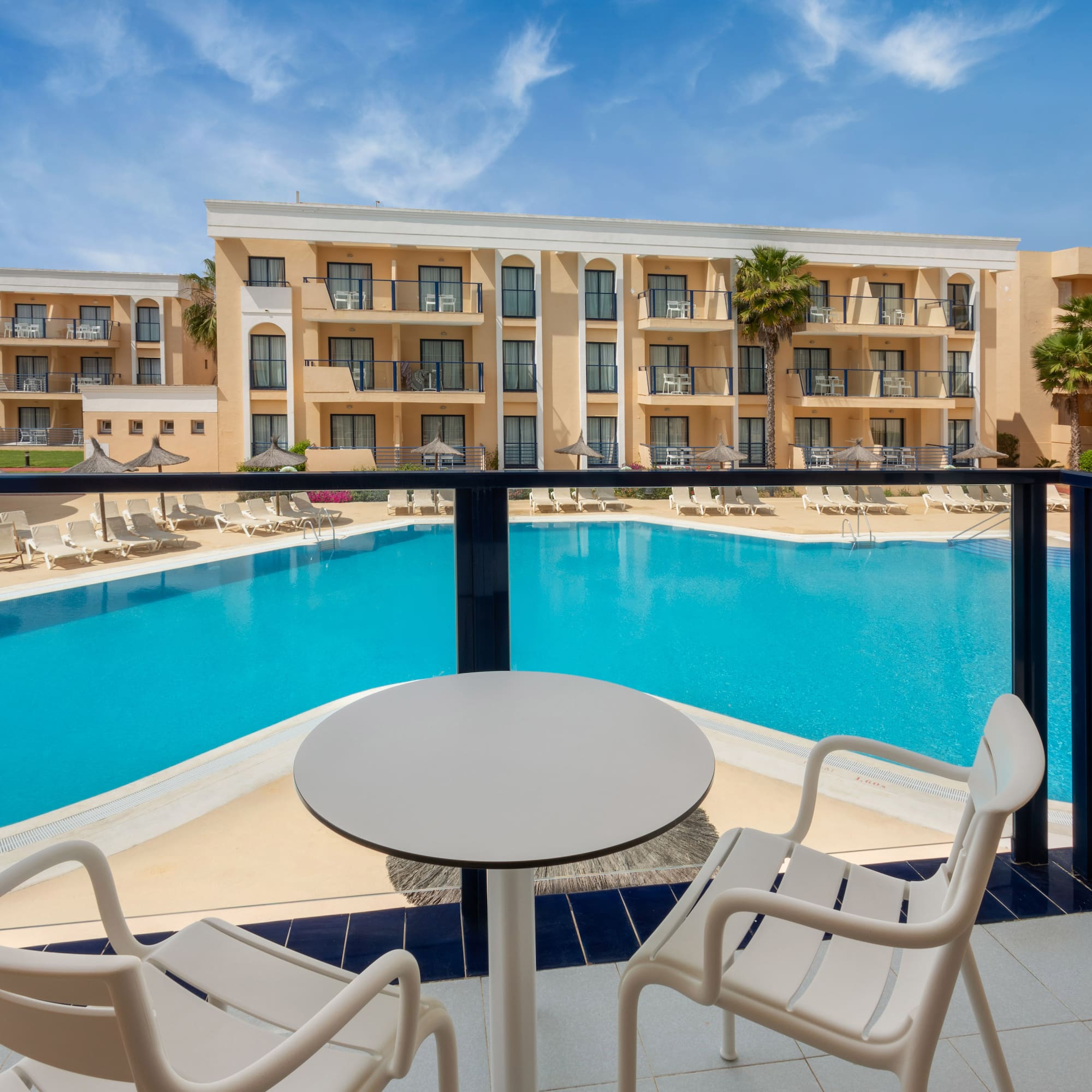 Resort hotel balcony view: large swimming pool, palm trees, sun loungers.