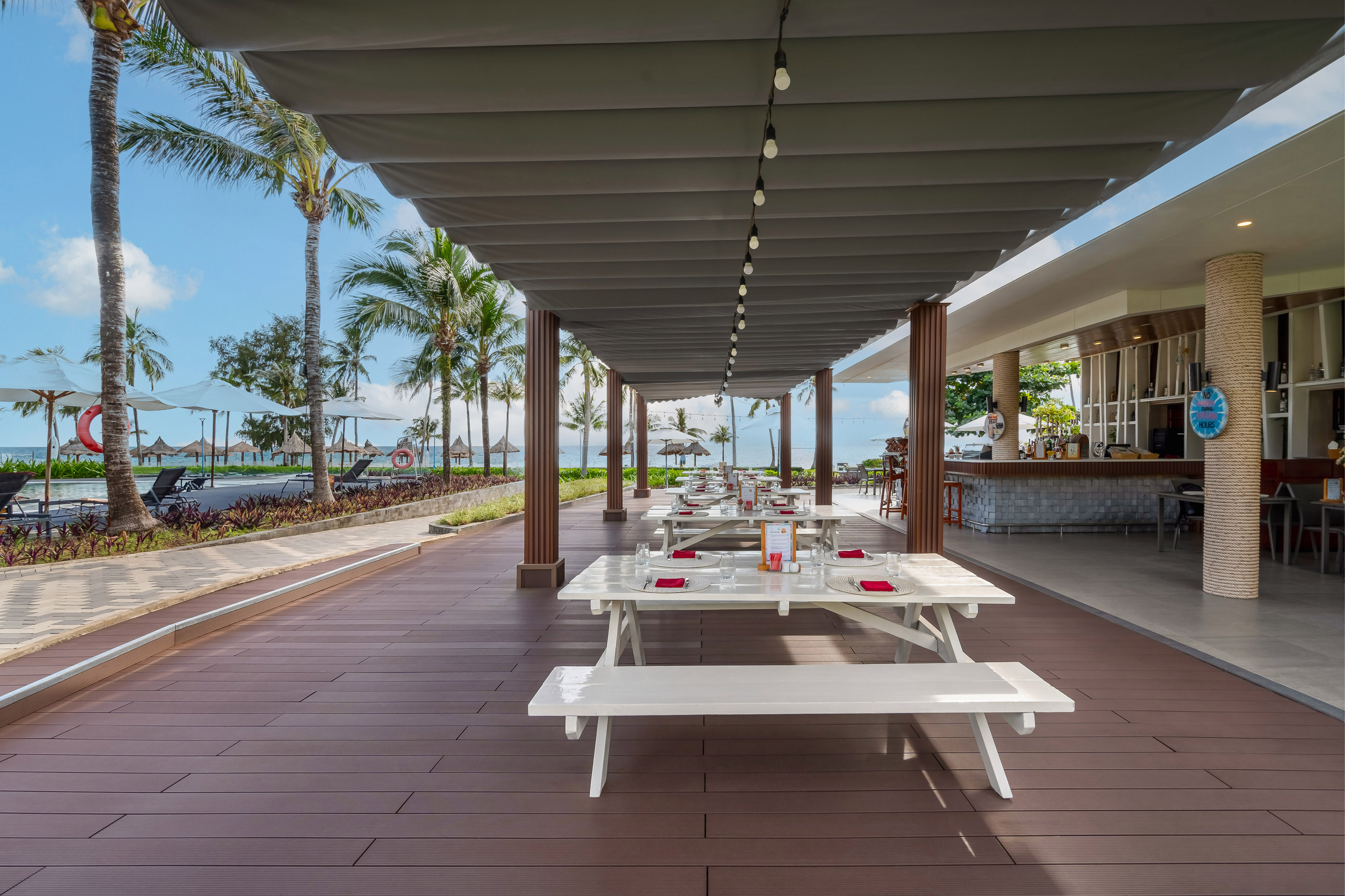 Beachfront restaurant patio with white picnic tables, palm trees, and ocean view.