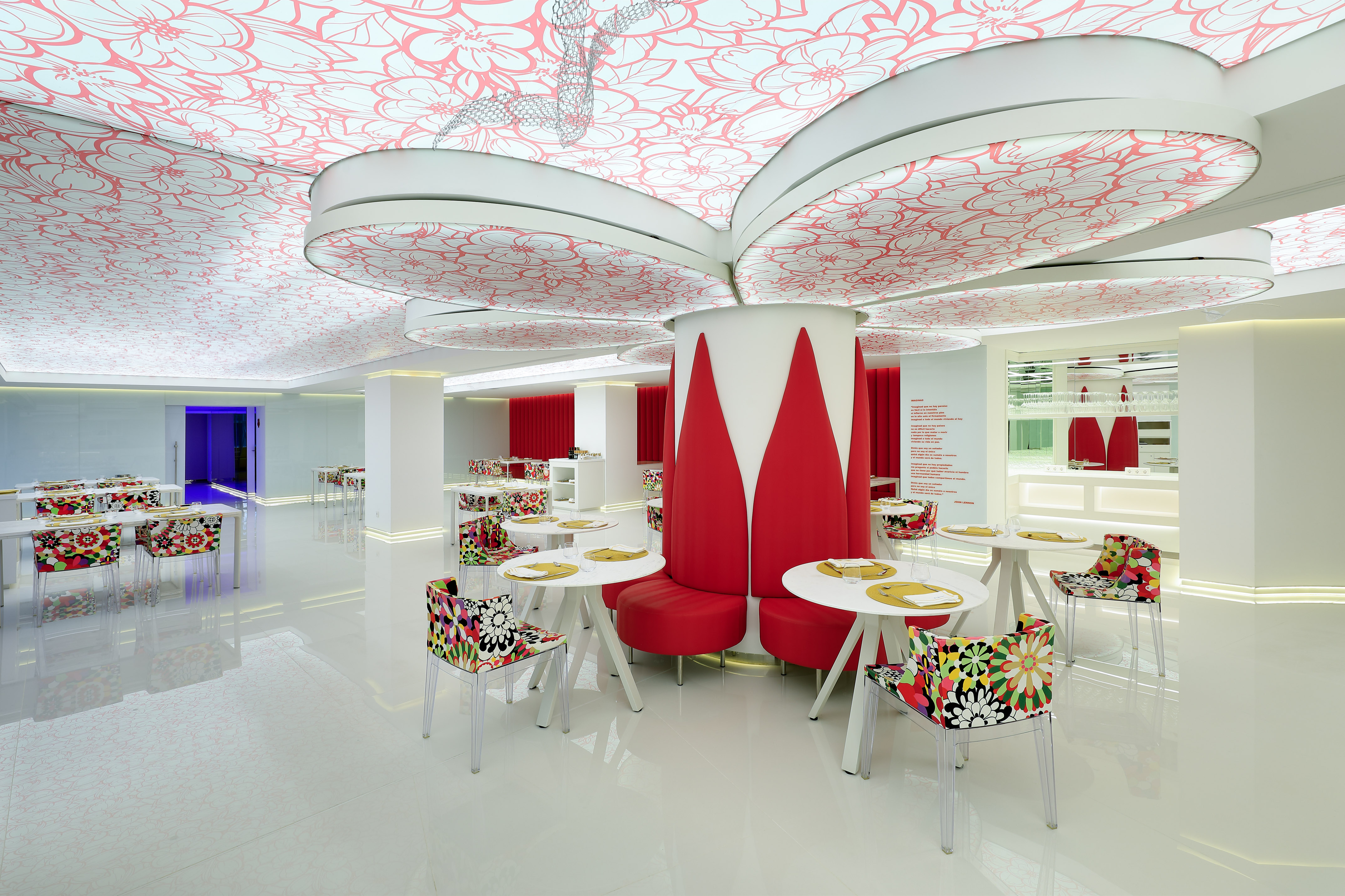 Modern restaurant with red floral ceiling, white decor, and colorful chairs.