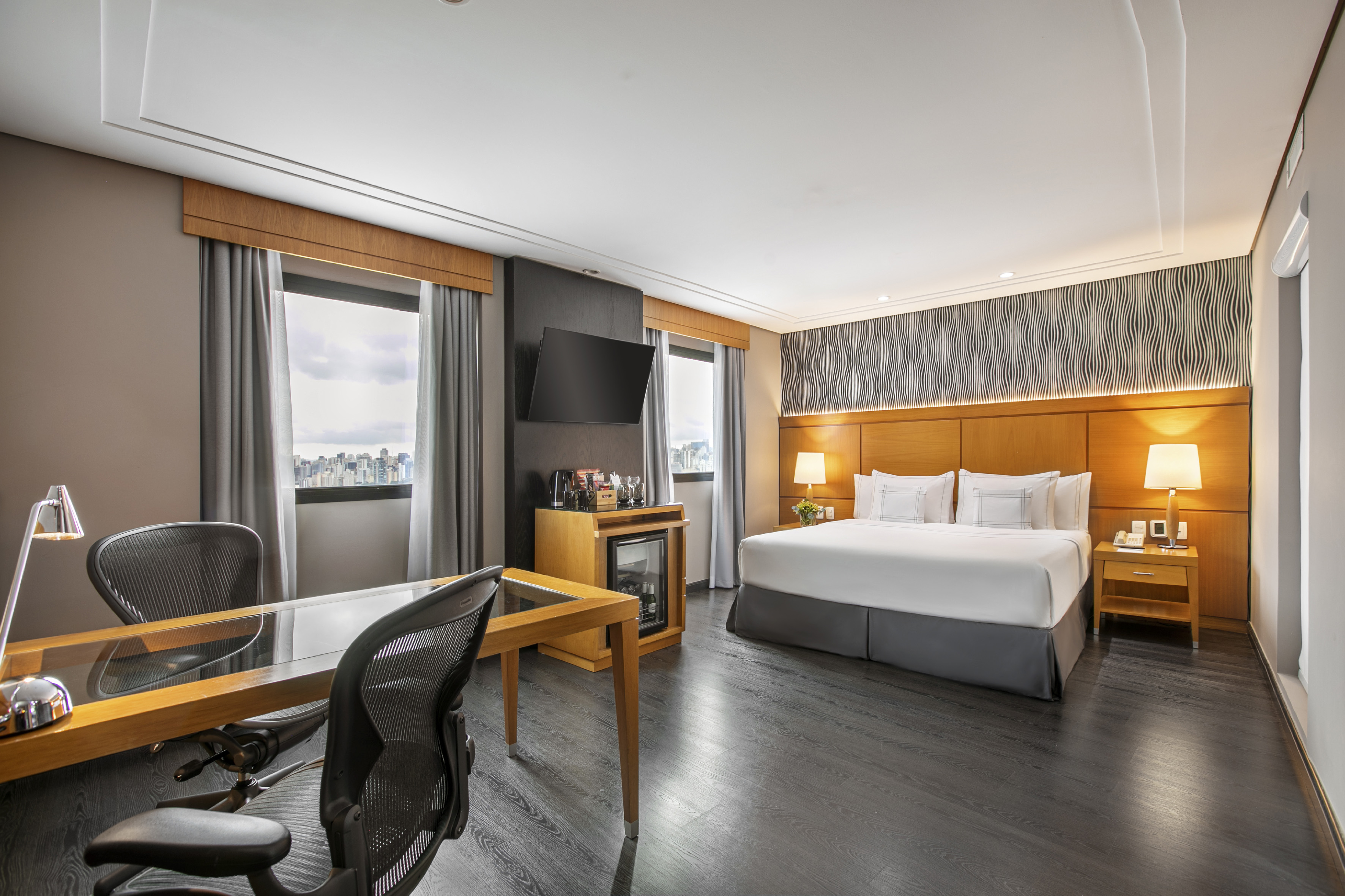 Modern hotel room with king bed, executive desk, city view, and flat-screen TV.