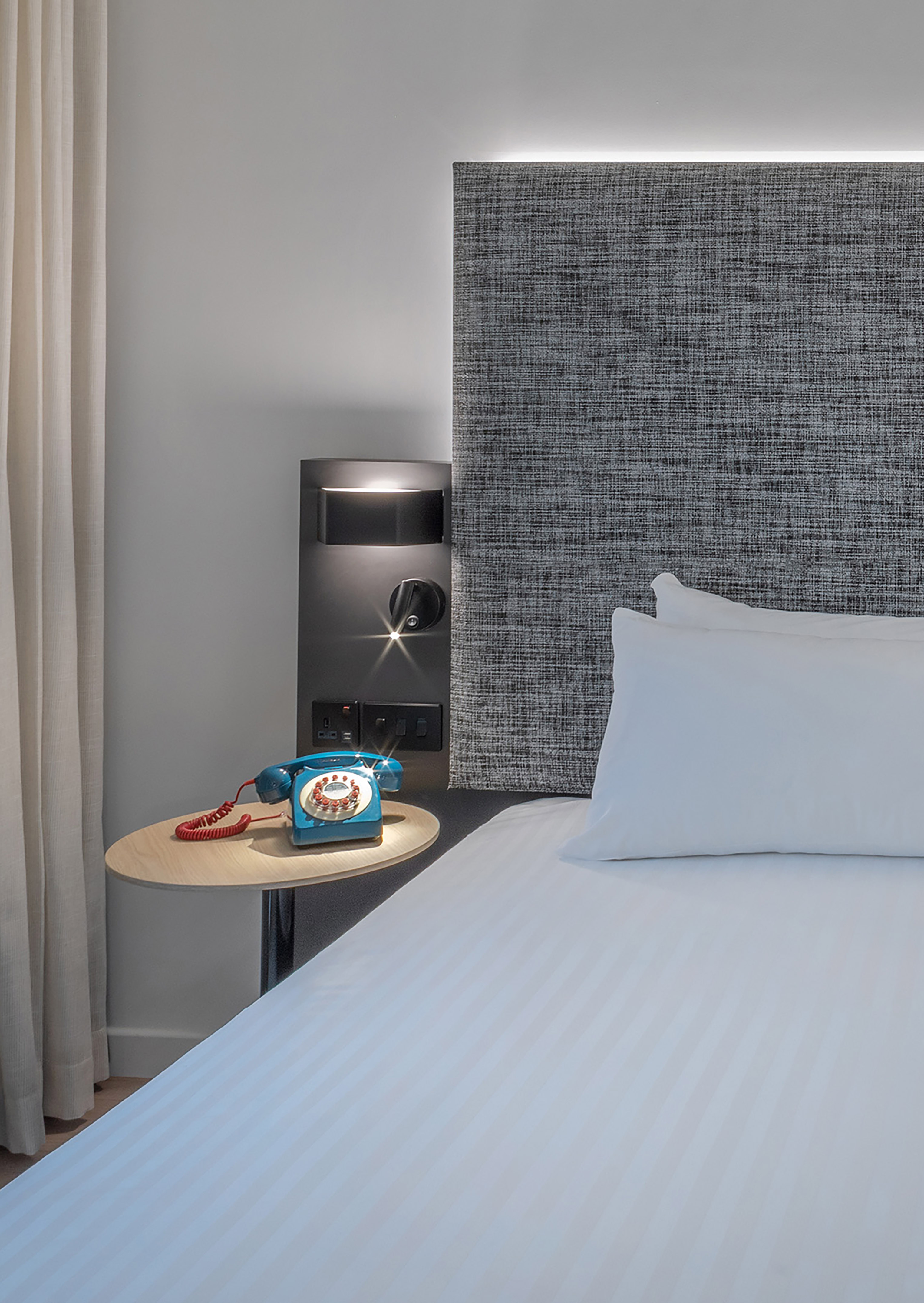Modern hotel bed, grey headboard, blue retro telephone on wooden bedside table.