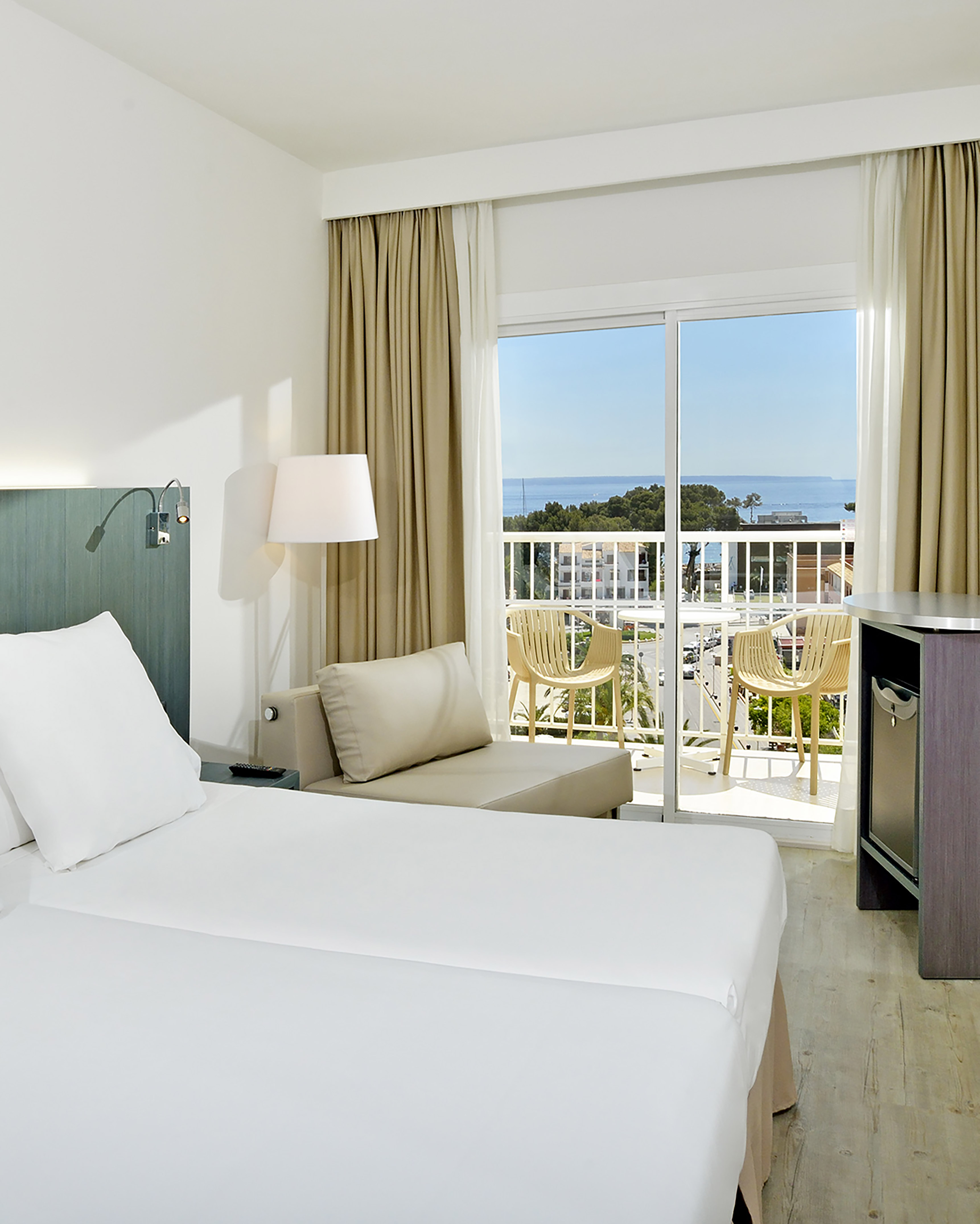 Modern hotel bedroom with white bed, chaise lounge, and sea view balcony.