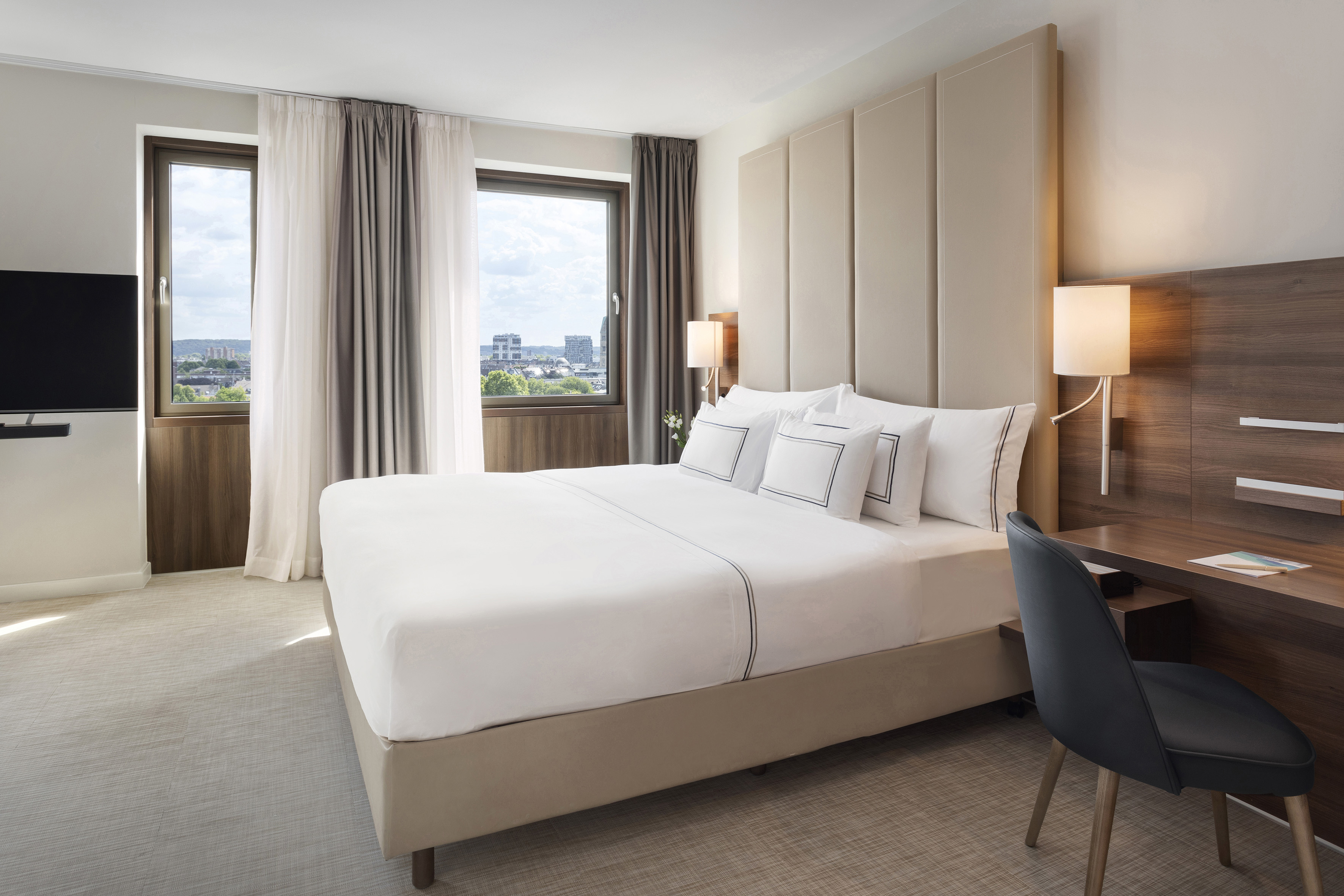 Modern hotel bedroom with large bed, upholstered headboard, city view windows, and desk.