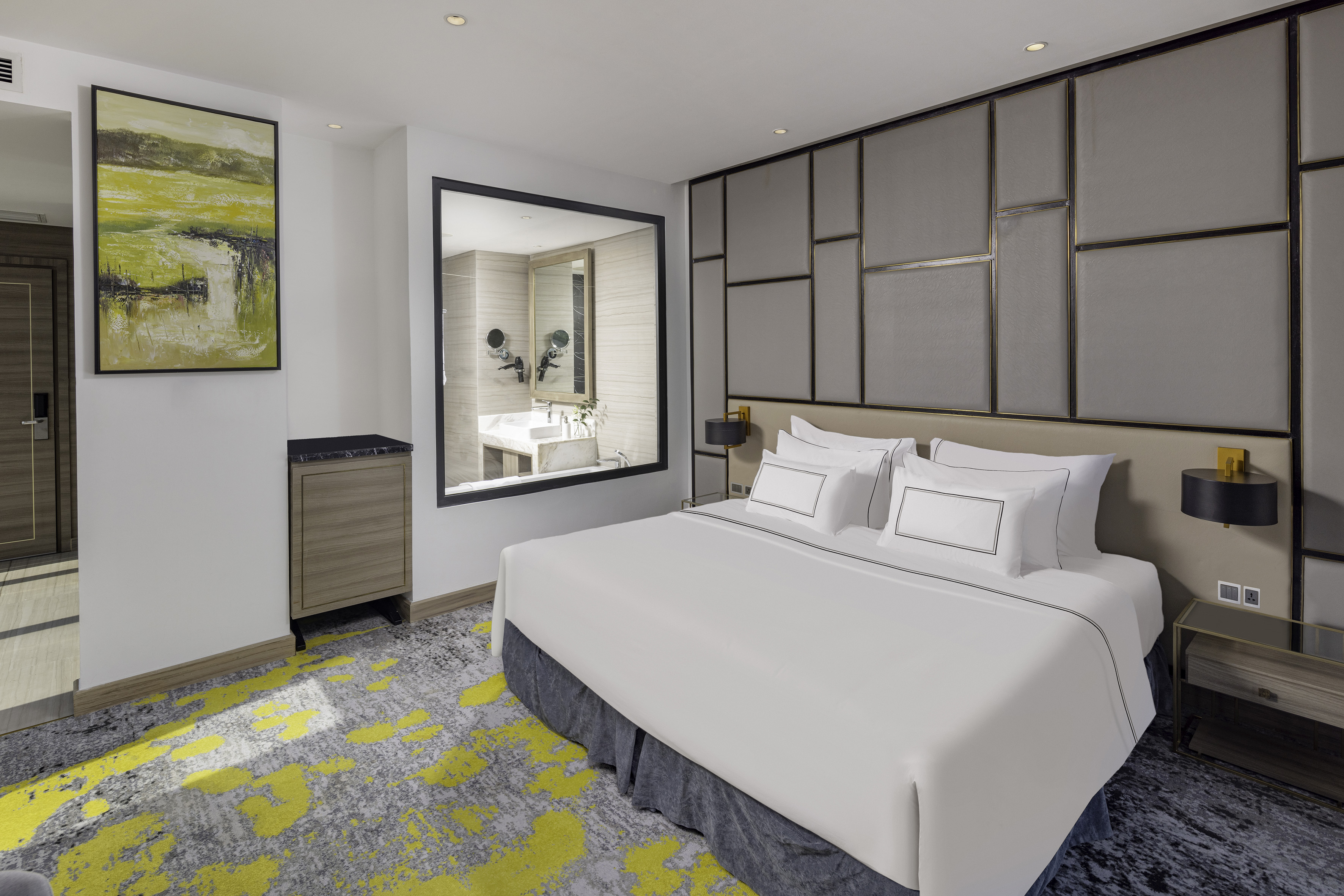 Modern hotel room with king bed, abstract yellow/grey carpet, and bathroom window.