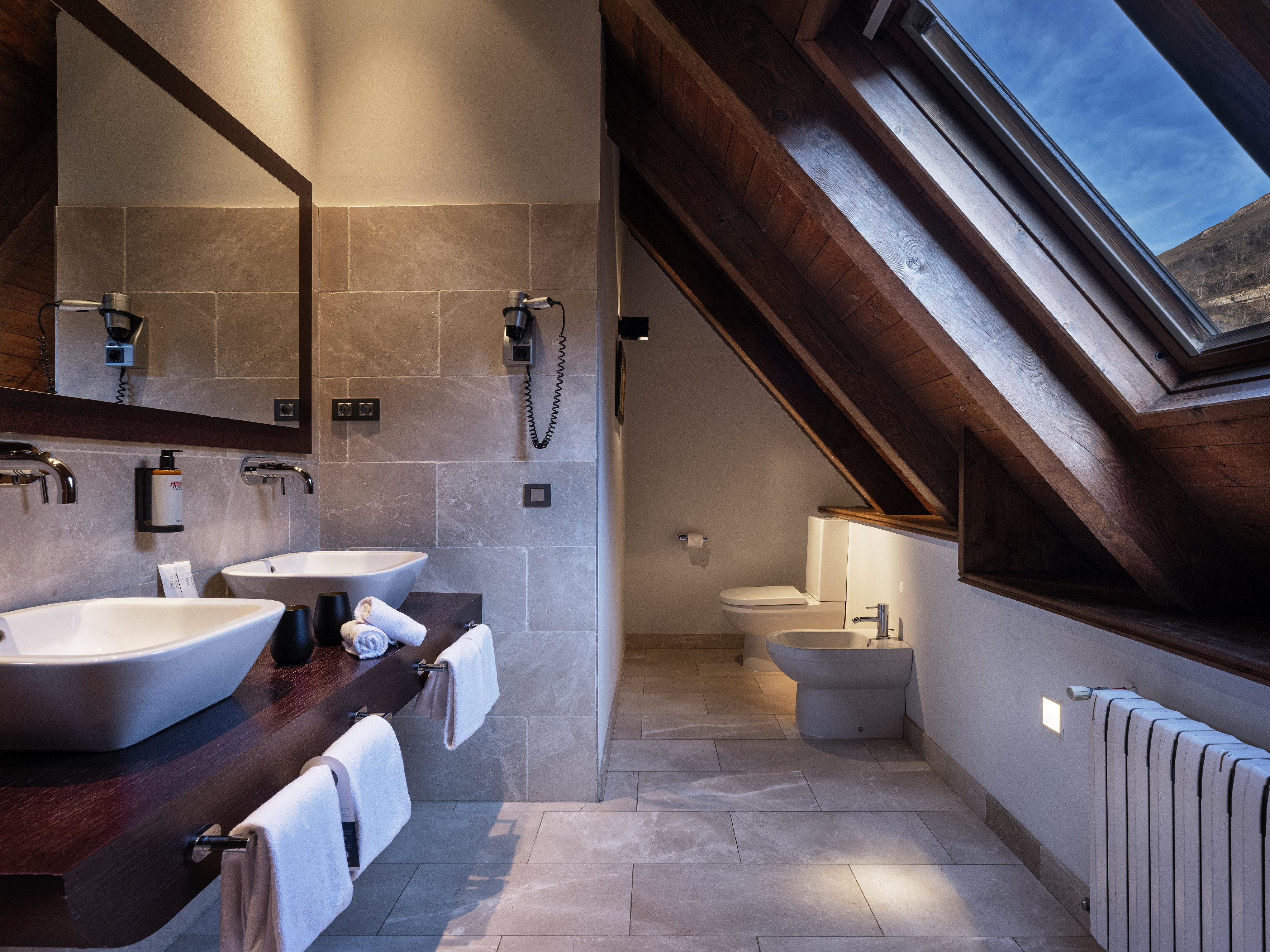 Modern attic bathroom with double vanity, wooden beams, and a bright skylight.