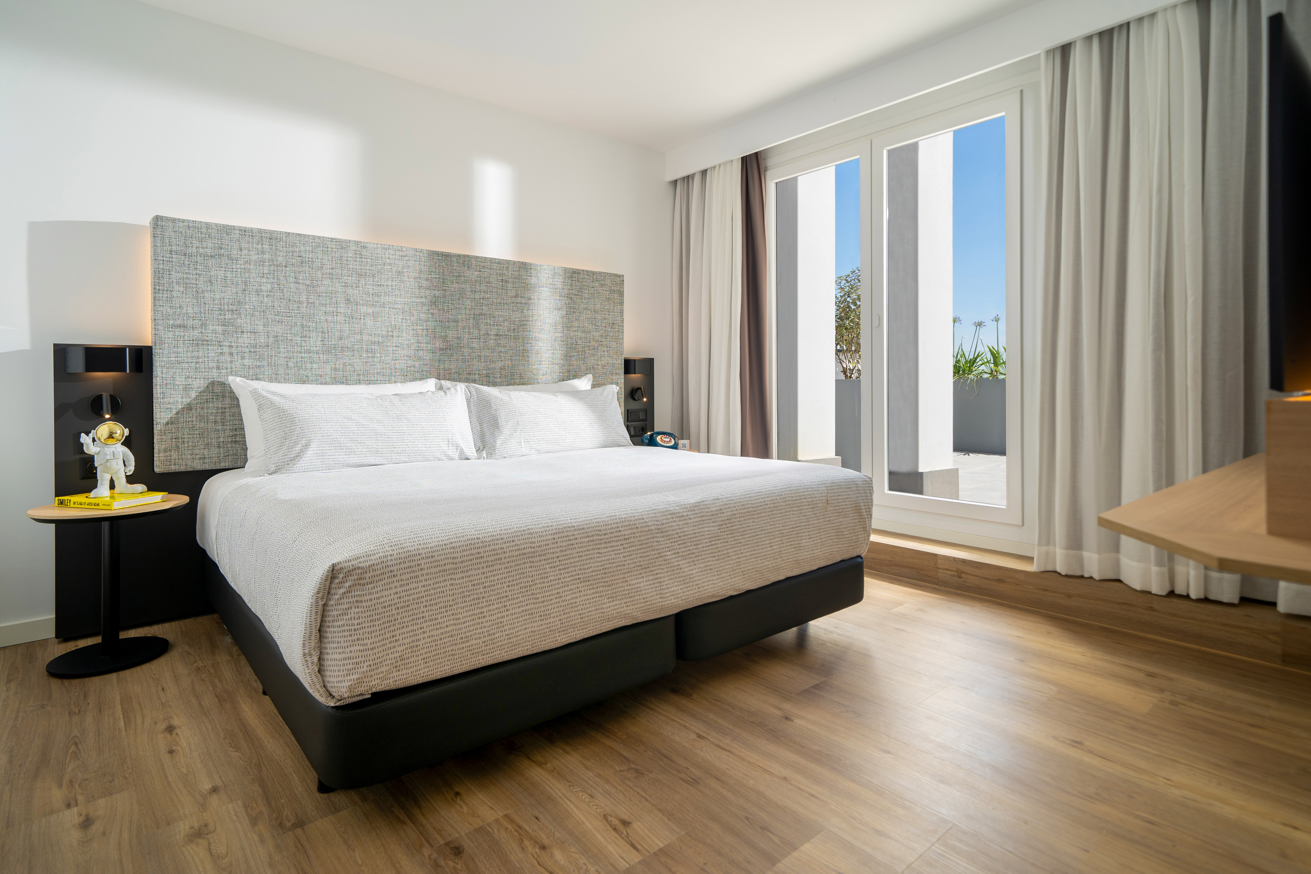 Modern hotel bedroom with queen bed, wood floor, and bright balcony access.