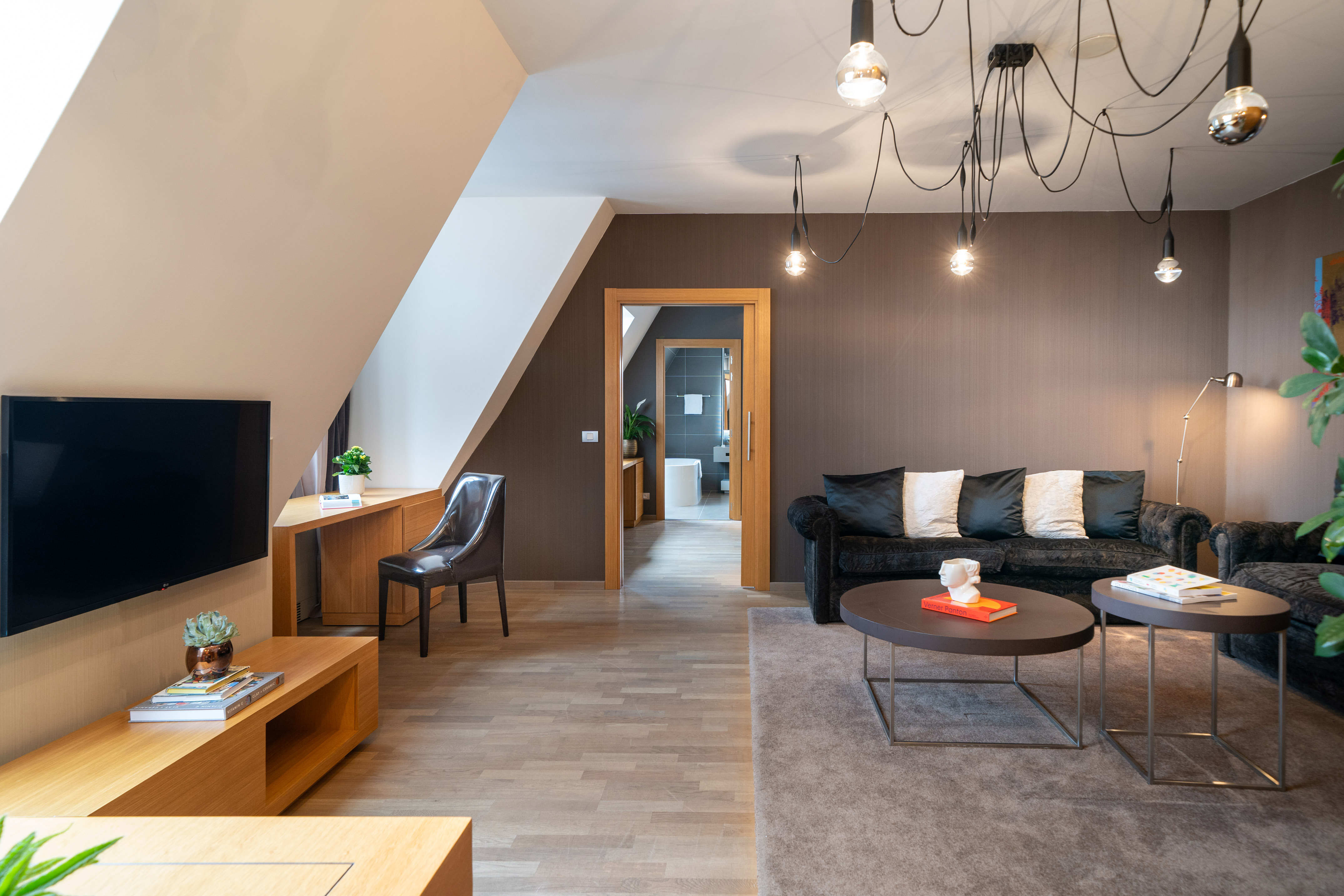 Modern attic living room with black sofa, TV, workspace, and bathroom entry.