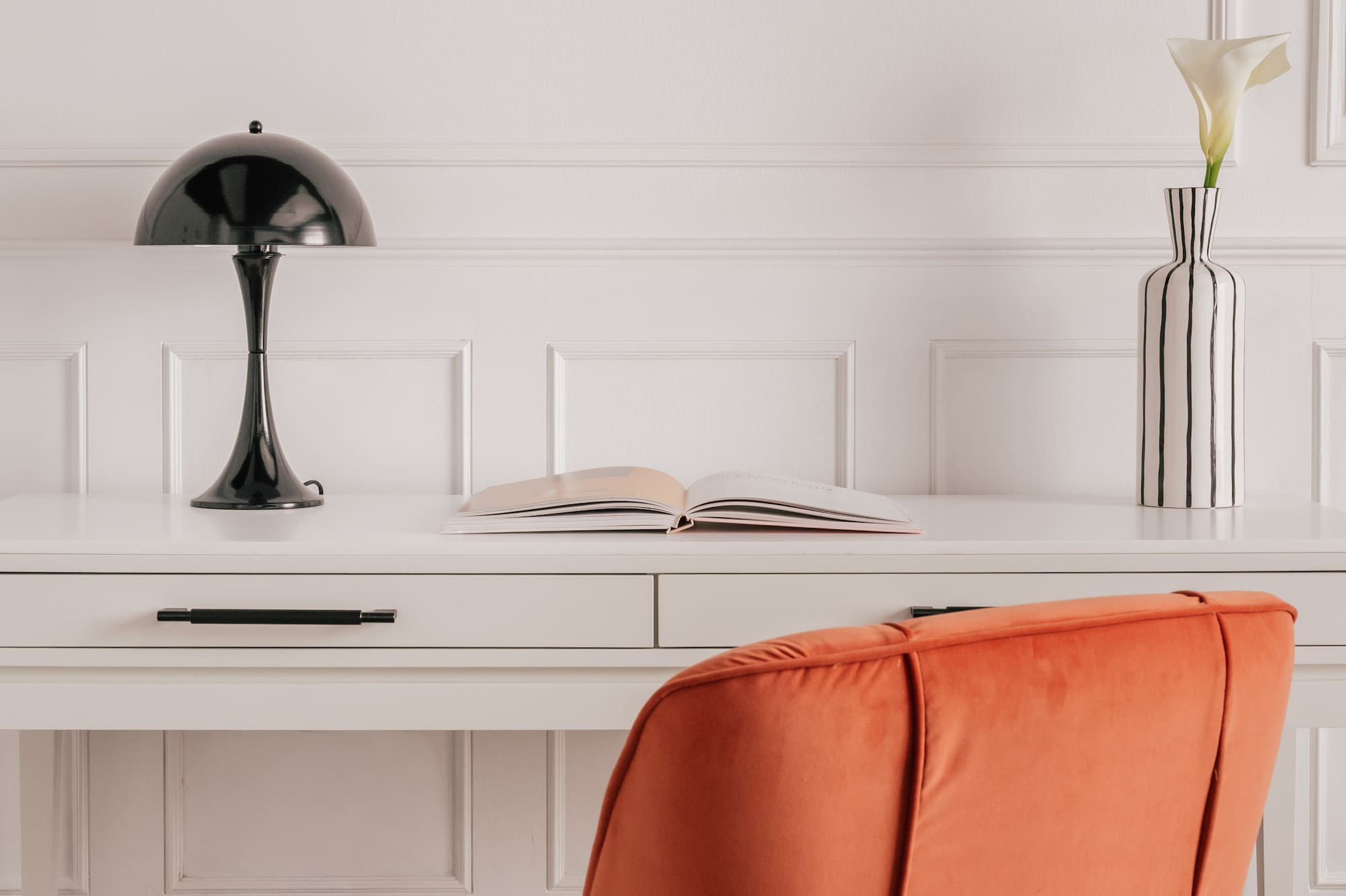 Modern home office with orange velvet chair, white desk, black lamp, striped vase.