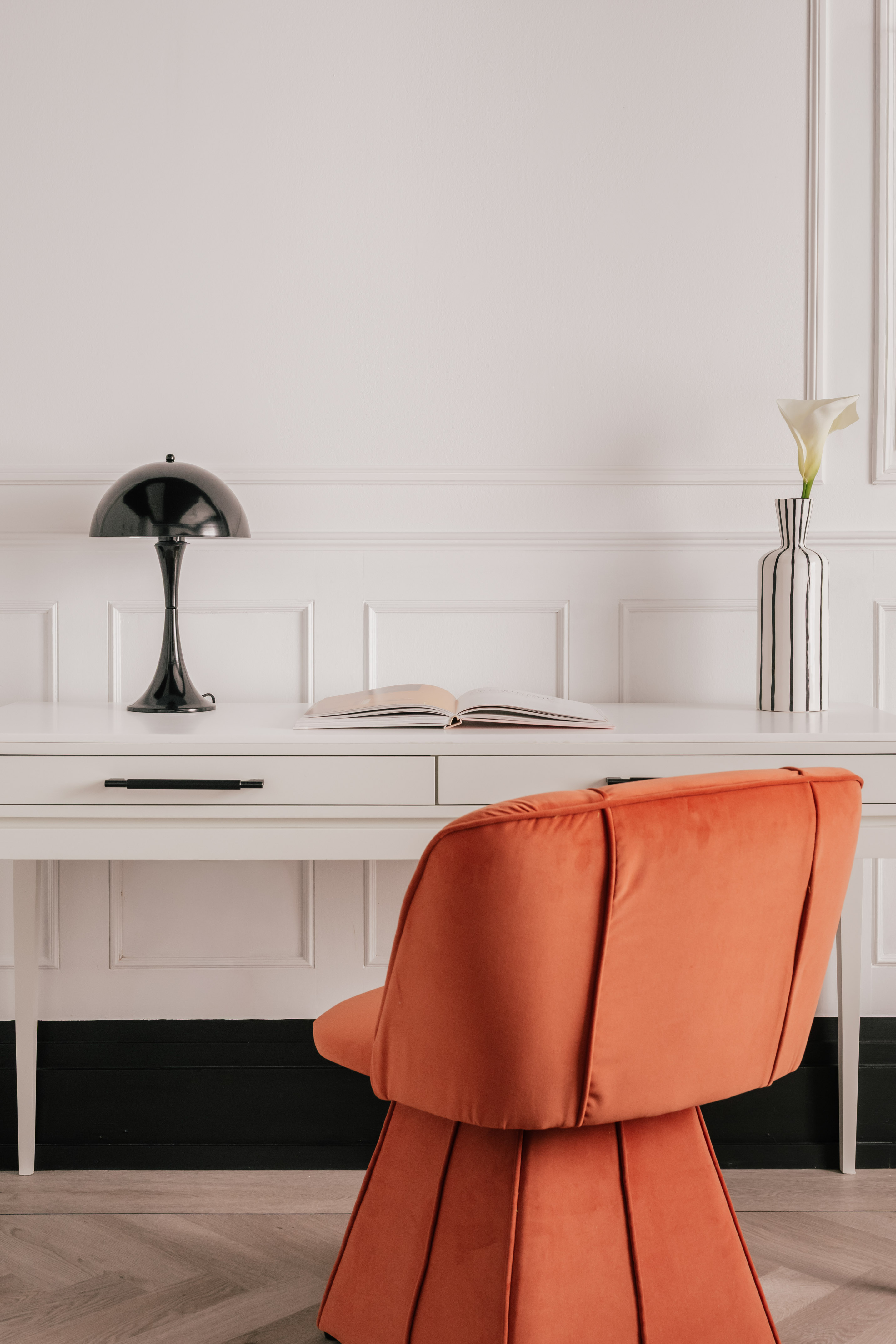 Modern home office with orange velvet chair, white desk, black lamp, striped vase.