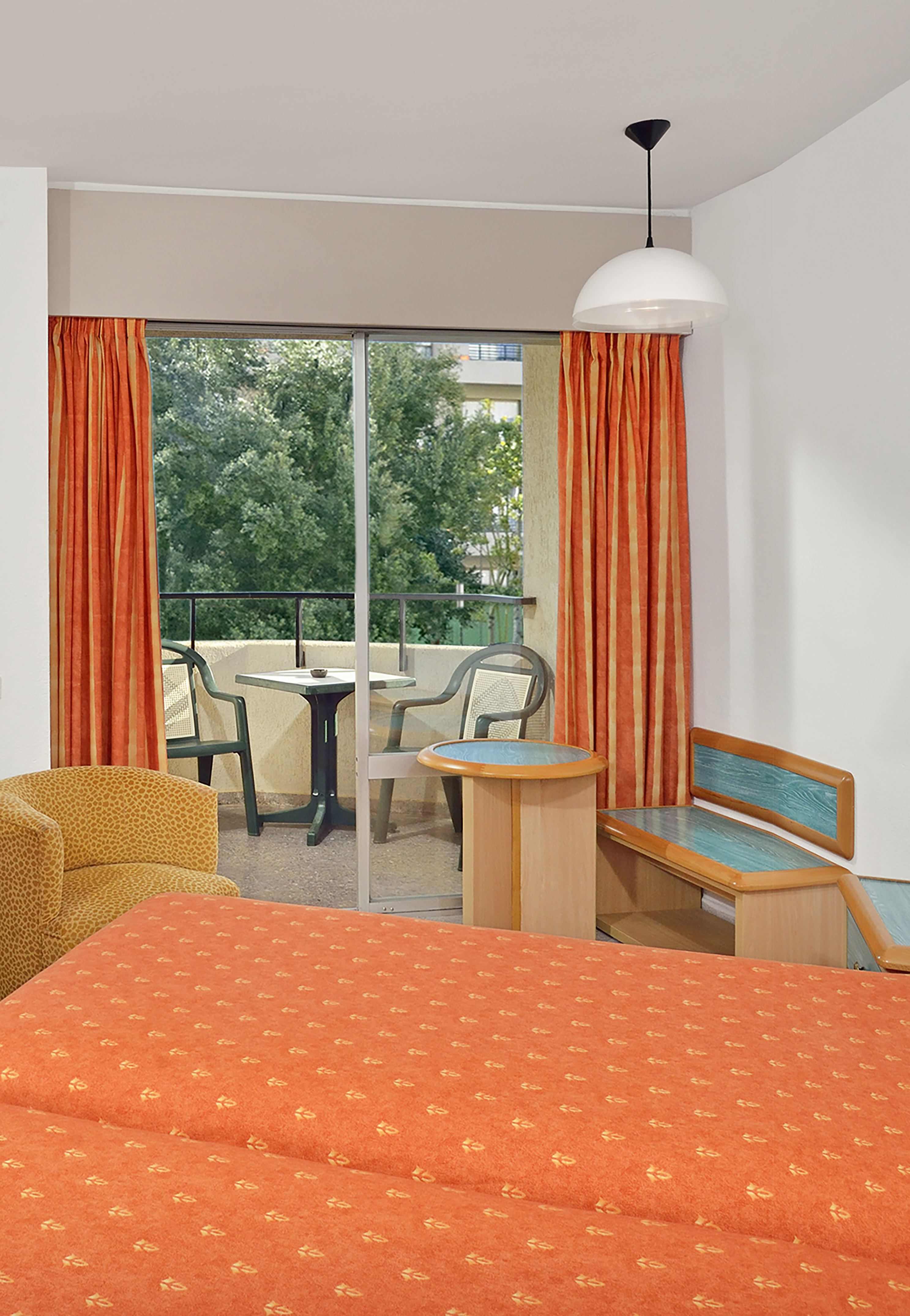 Hotel bedroom with vibrant orange bed, balcony with seating, and green trees.