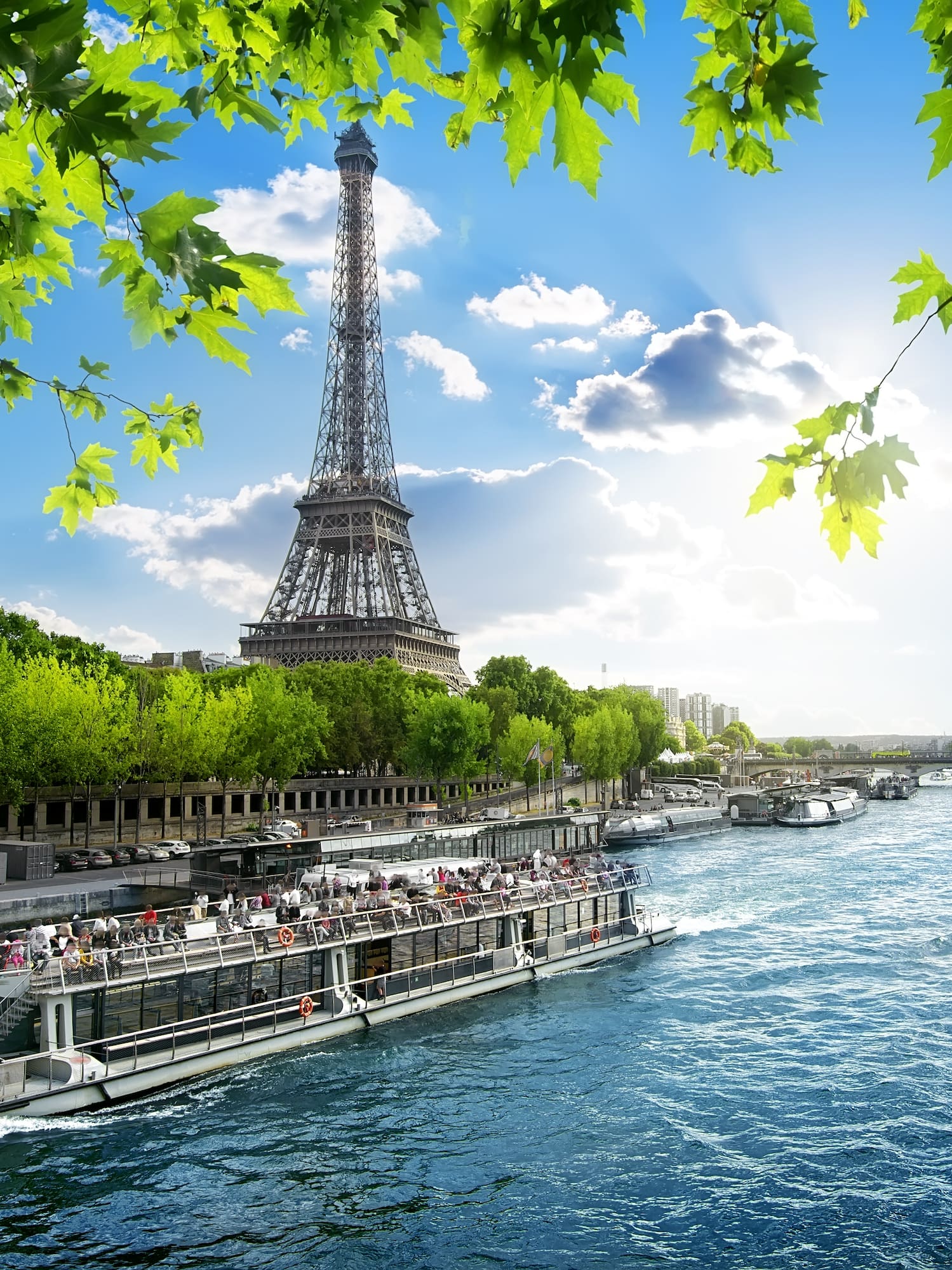 Eiffel Tower over Seine River with tour boat, green leaves, sunny Paris.