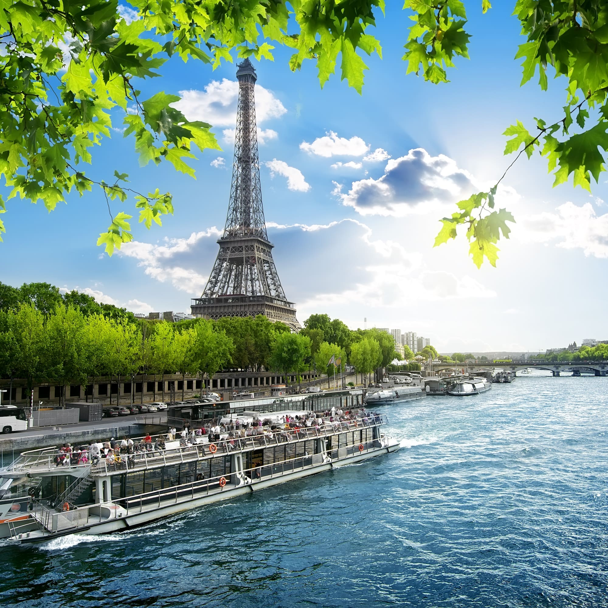 Eiffel Tower over Seine River with tour boat, green leaves, sunny Paris.